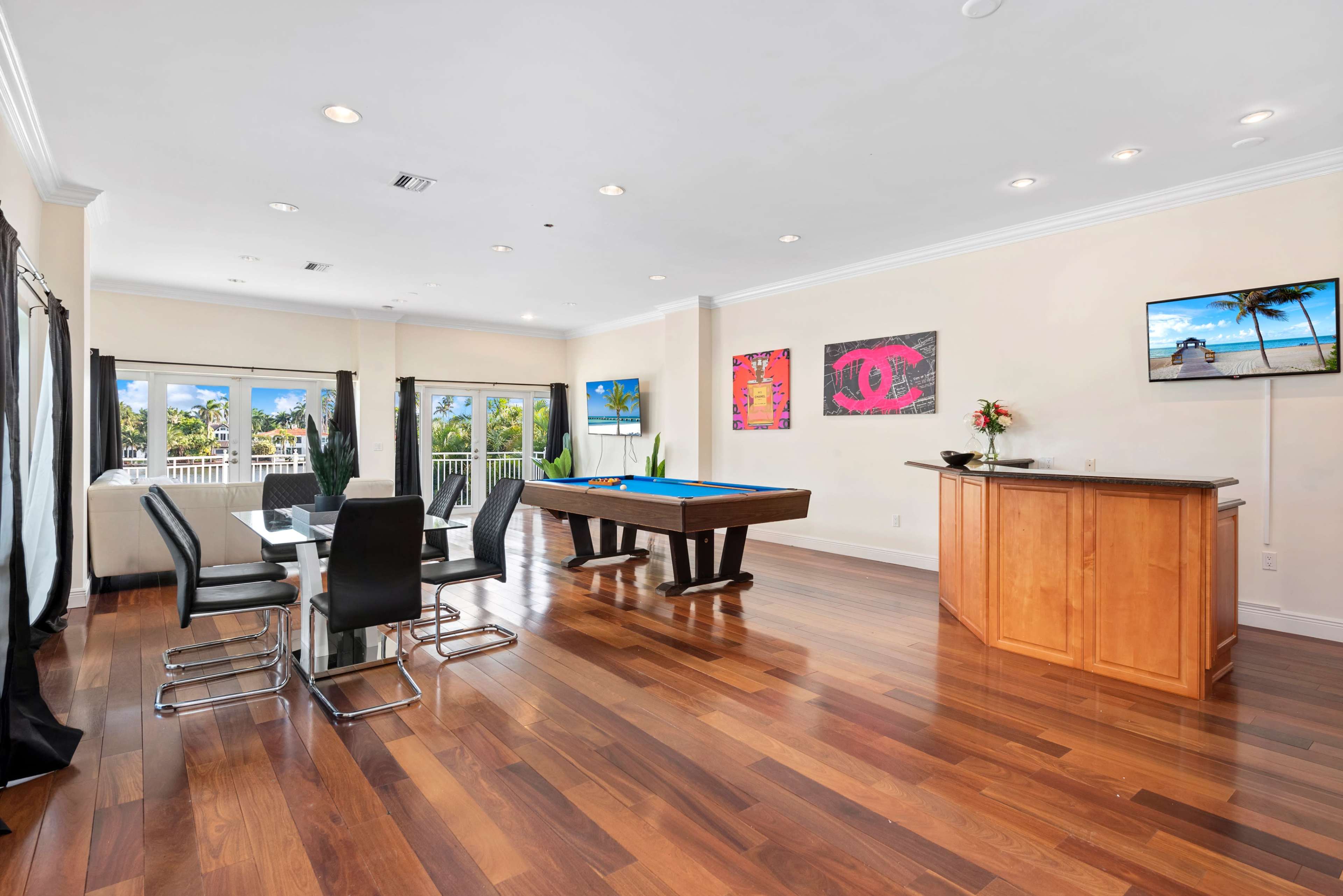 The image shows a modern game room featuring a pool table, a dining table with black chairs, and flat-screen TVs on the walls, all situated on polished wooden floors.