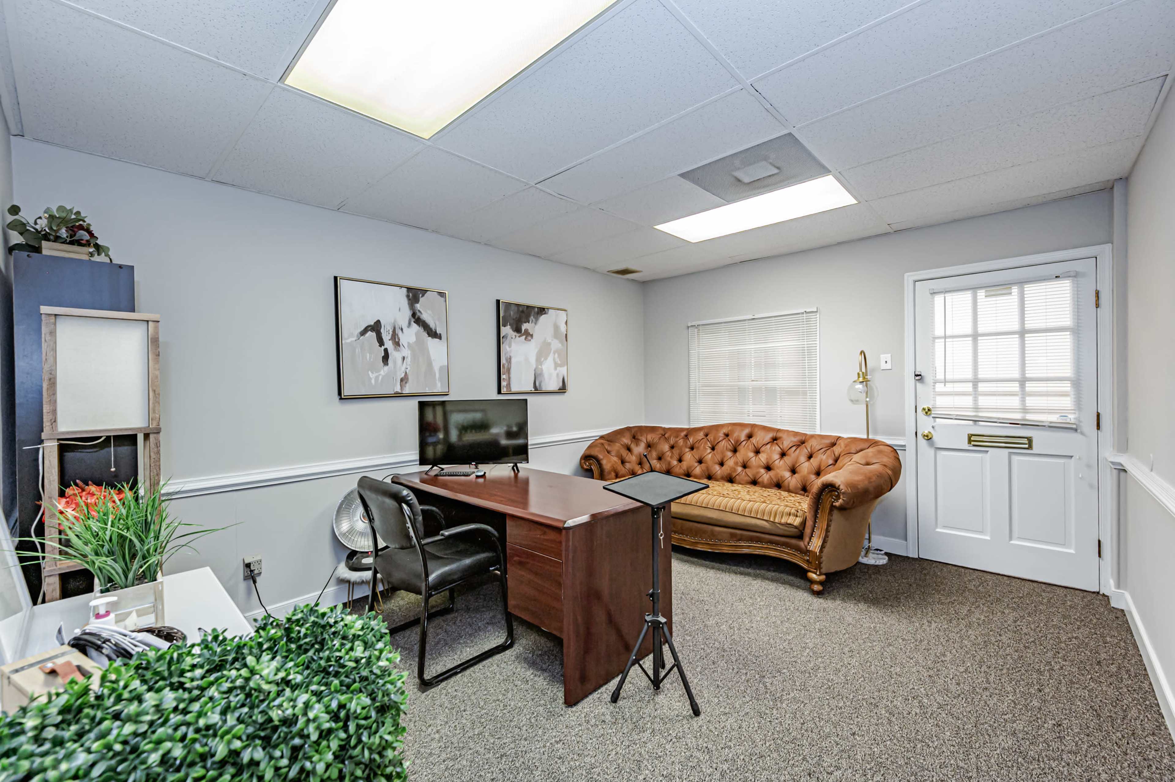 A small office space with a brown tufted couch, a wooden desk, a television, and door leading outside.