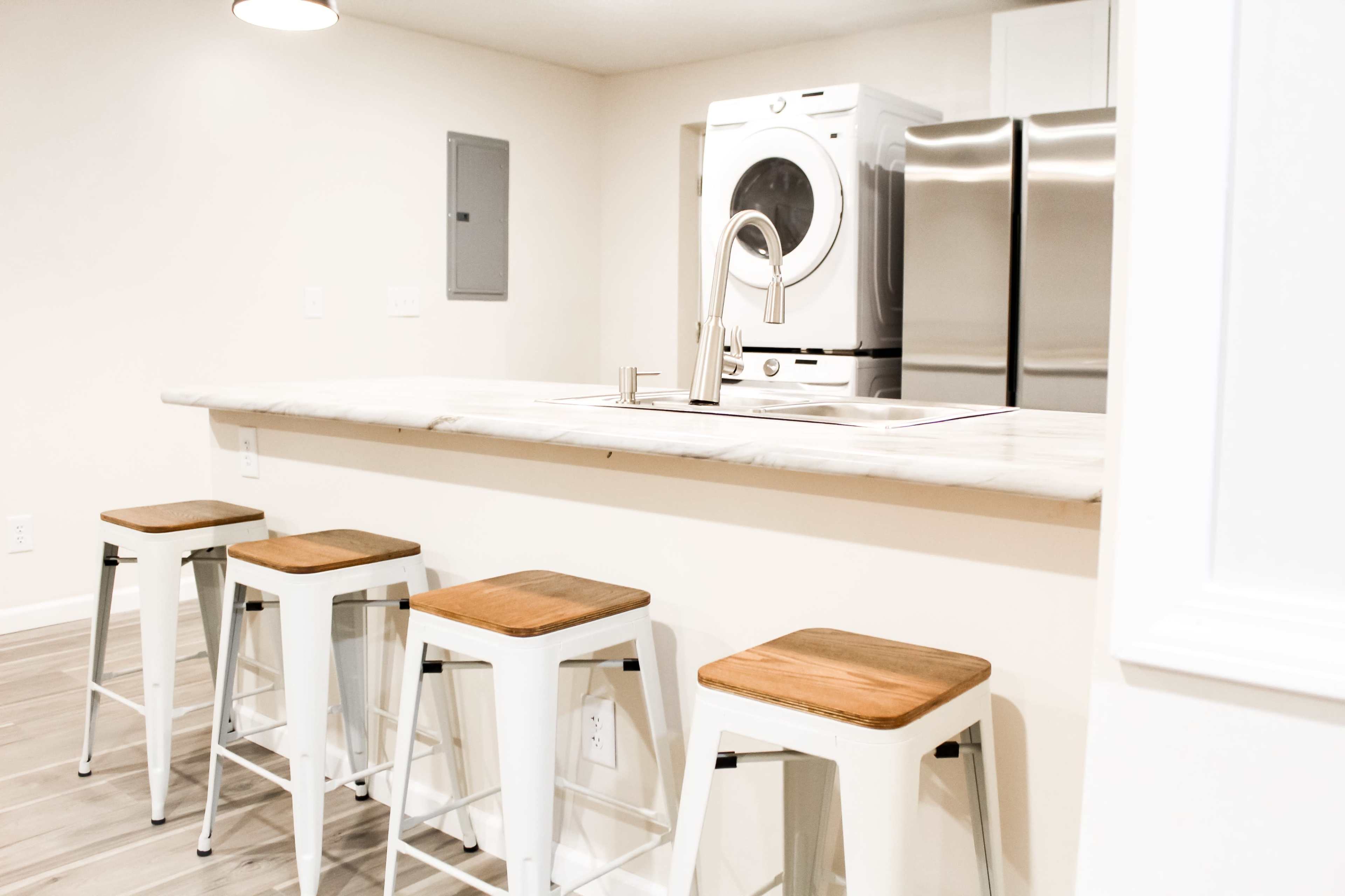 The image shows a modern kitchen with a marble-topped bar and four metal stools, featuring a washer and dryer unit and stainless steel appliances in the background.