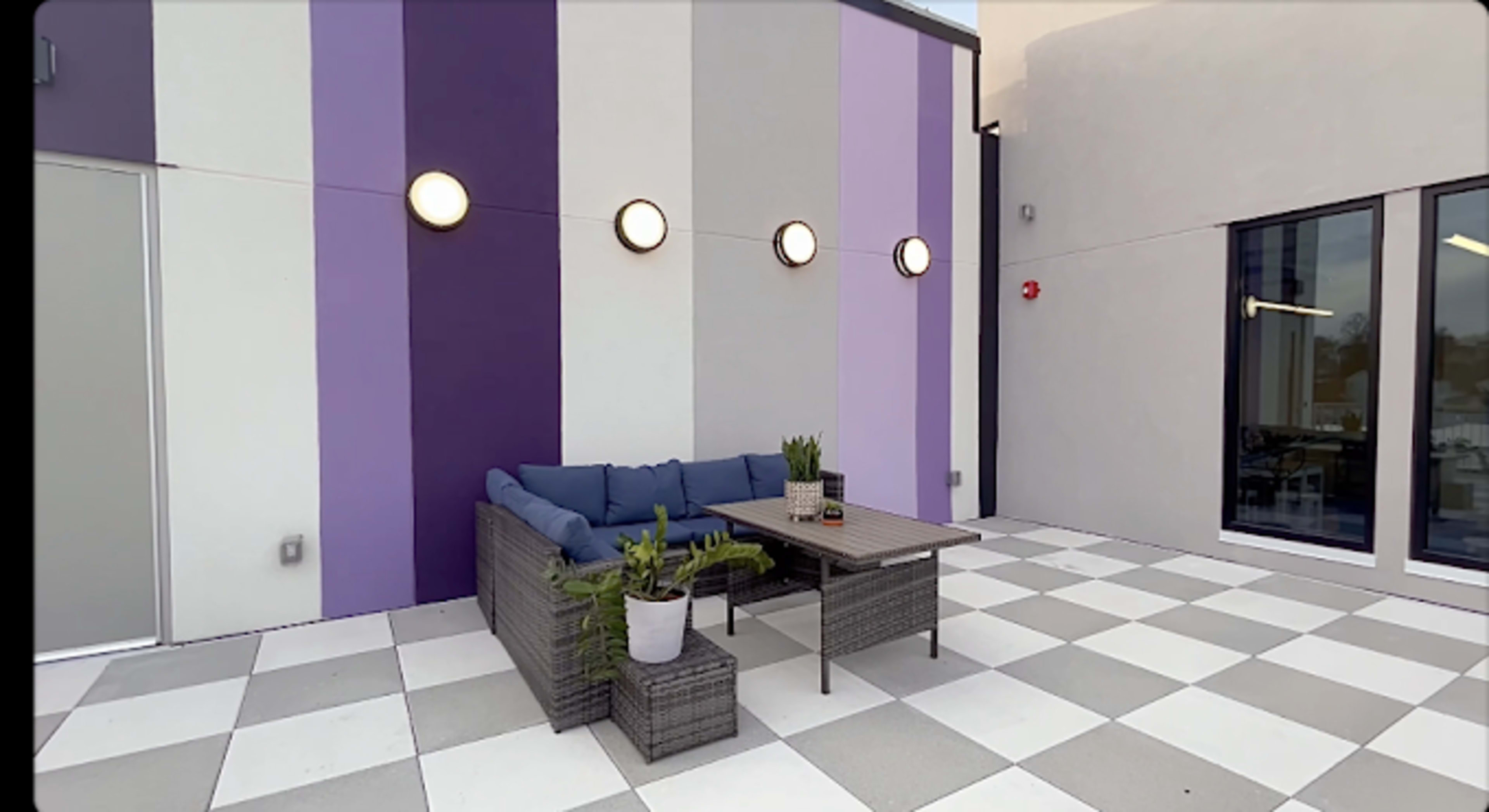 The image shows a modern outdoor seating area with a sectional sofa, table, and potted plants against a wall featuring purple and white stripes.