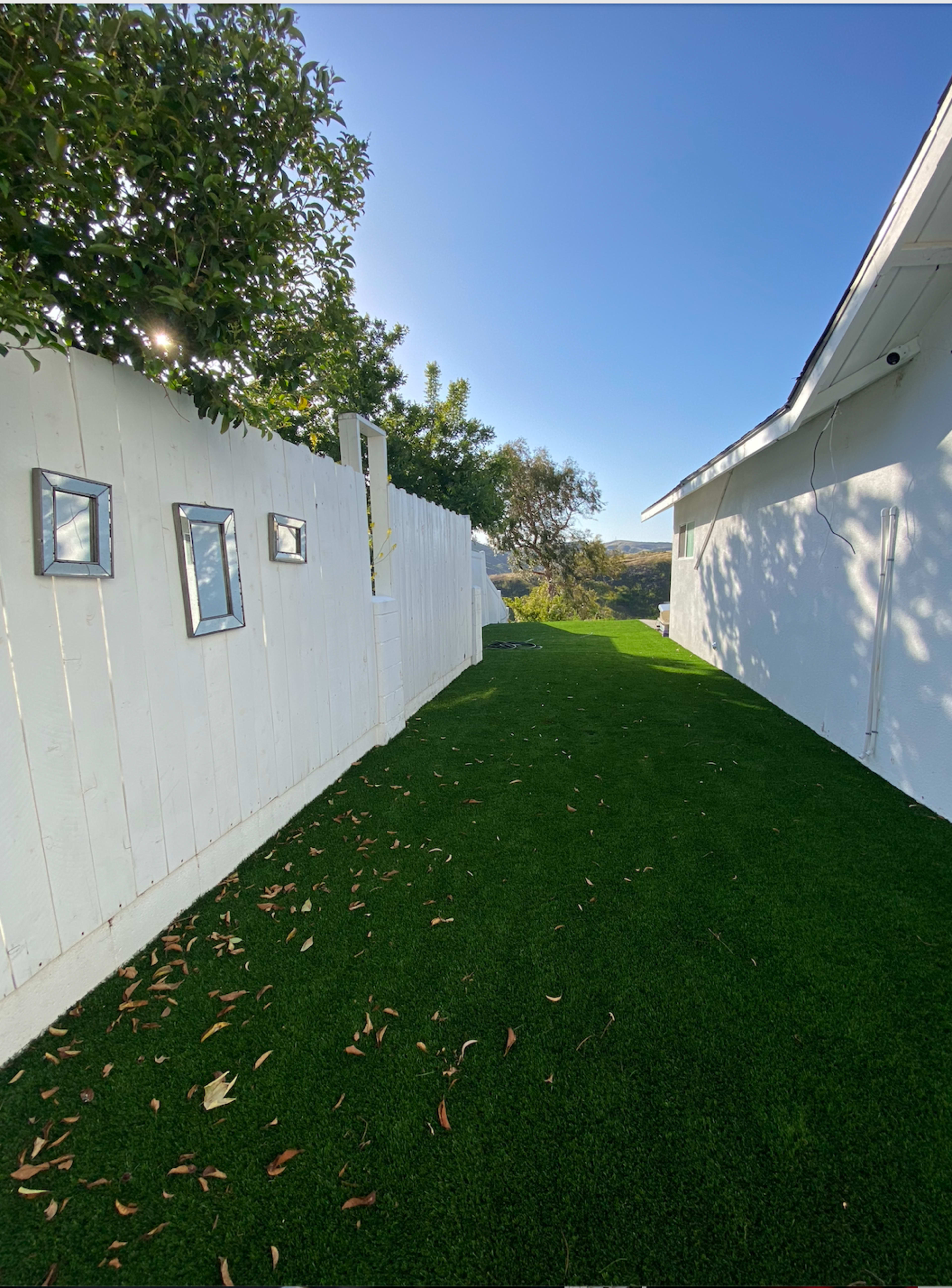 A narrow pathway lined with white fences and artificial grass leads toward a house on a clear day.