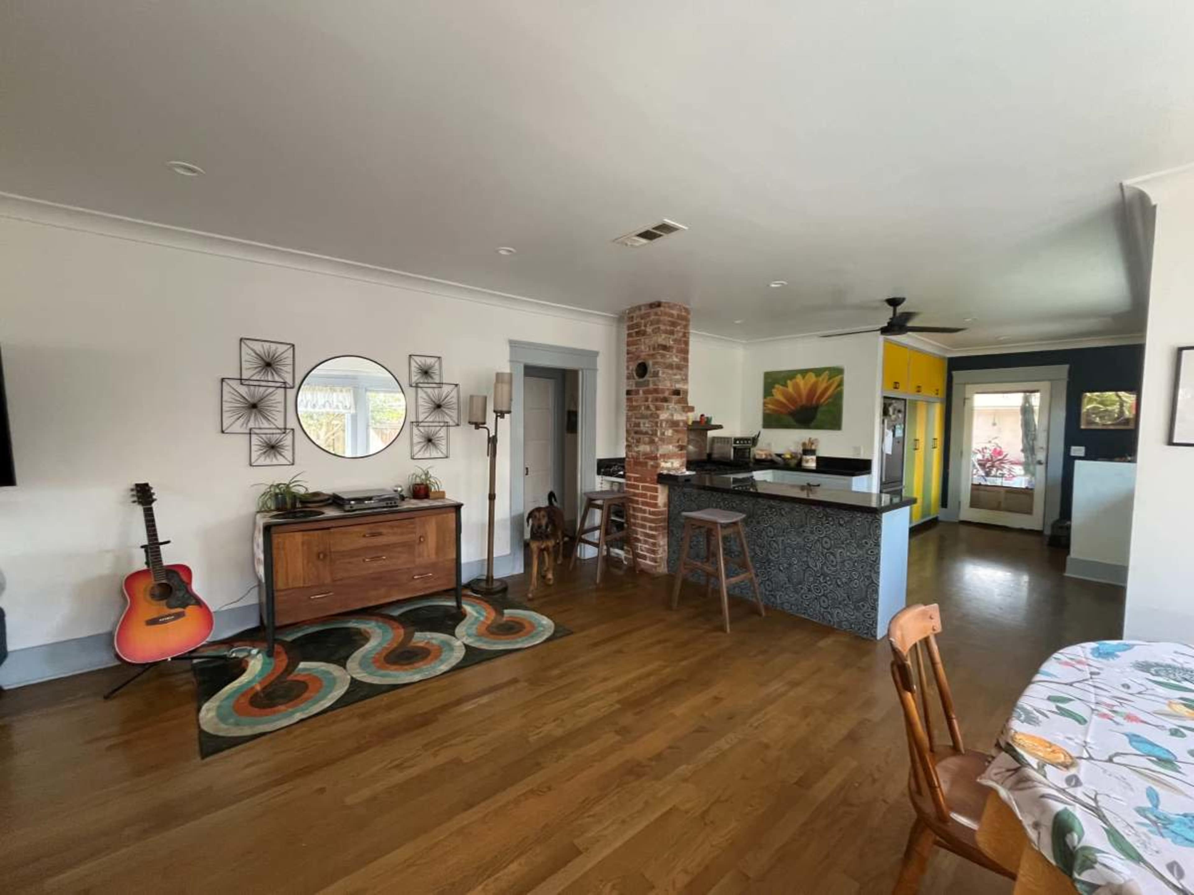 The image shows a well-lit open-concept living space featuring hardwood floors, a dining table, a kitchen with a bold yellow wall, and a guitar propped near a console table.