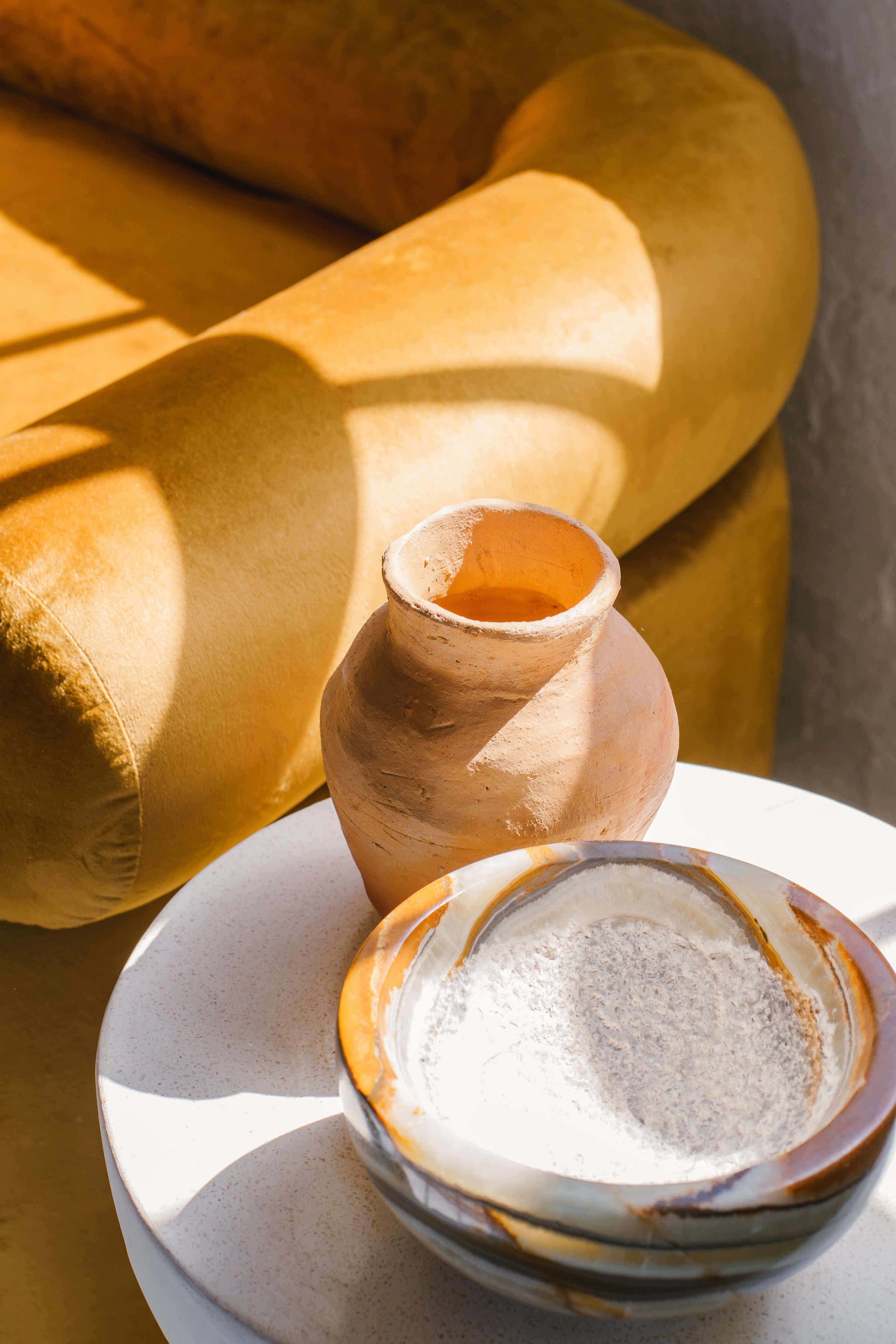 A clay pot and a bowl of powder sit on a round table beside a yellow velvet sofa.
