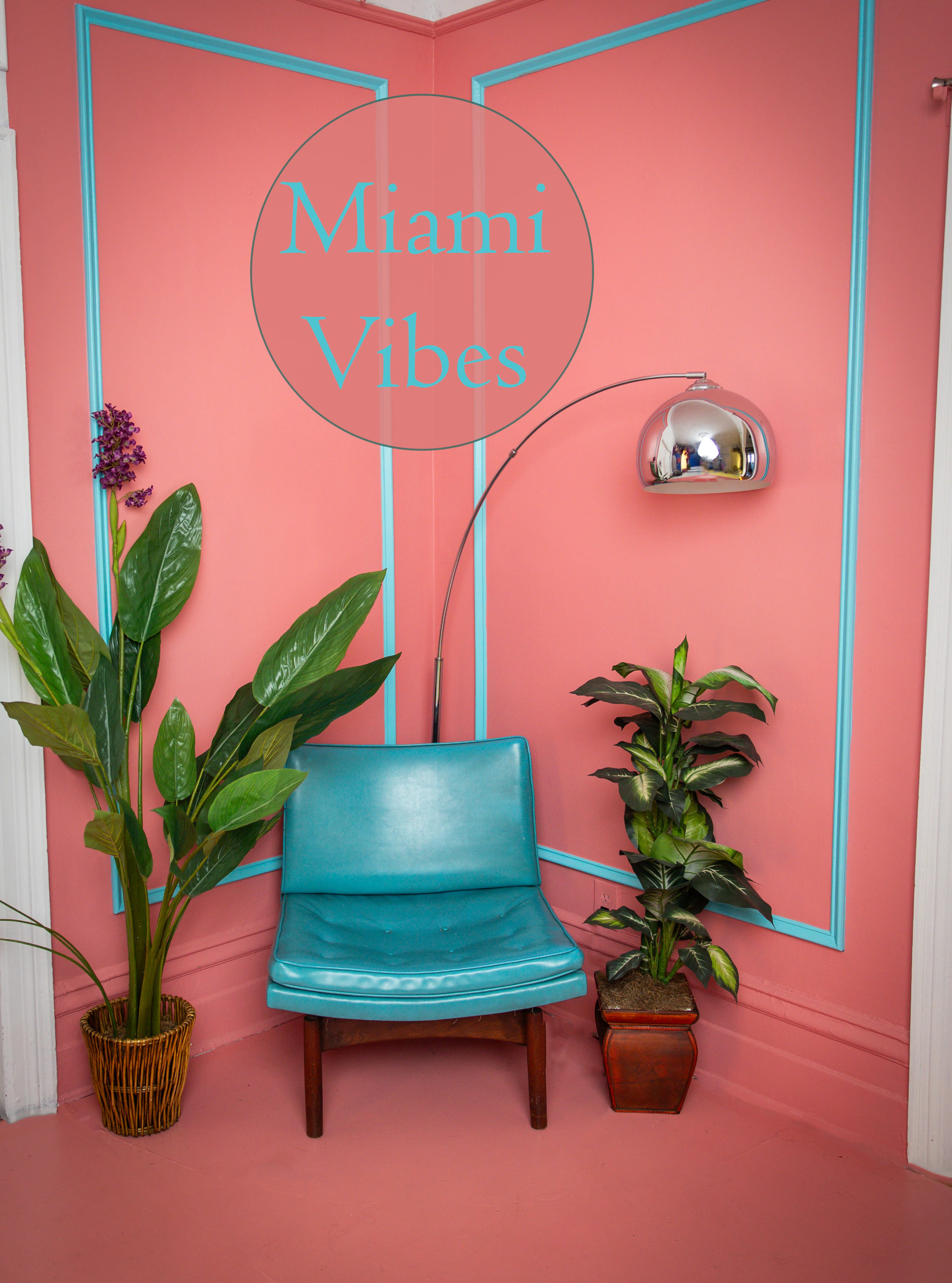 The image features a teal chair positioned against a pink wall with blue trim, accompanied by decorative plants and a modern arc floor lamp.