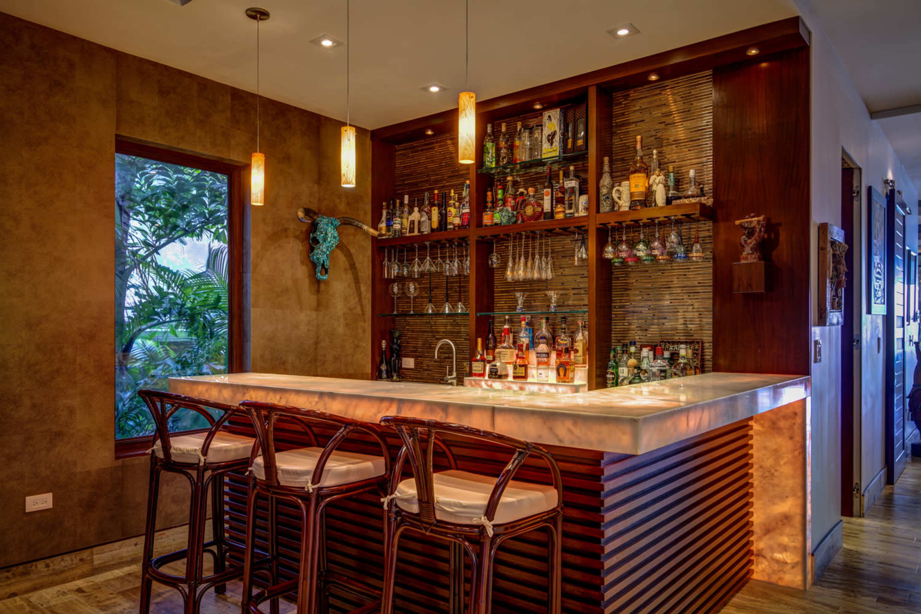 The image shows a modern bar area with a wooden counter, several bottles of alcohol displayed on shelves, and three bar stools in front.