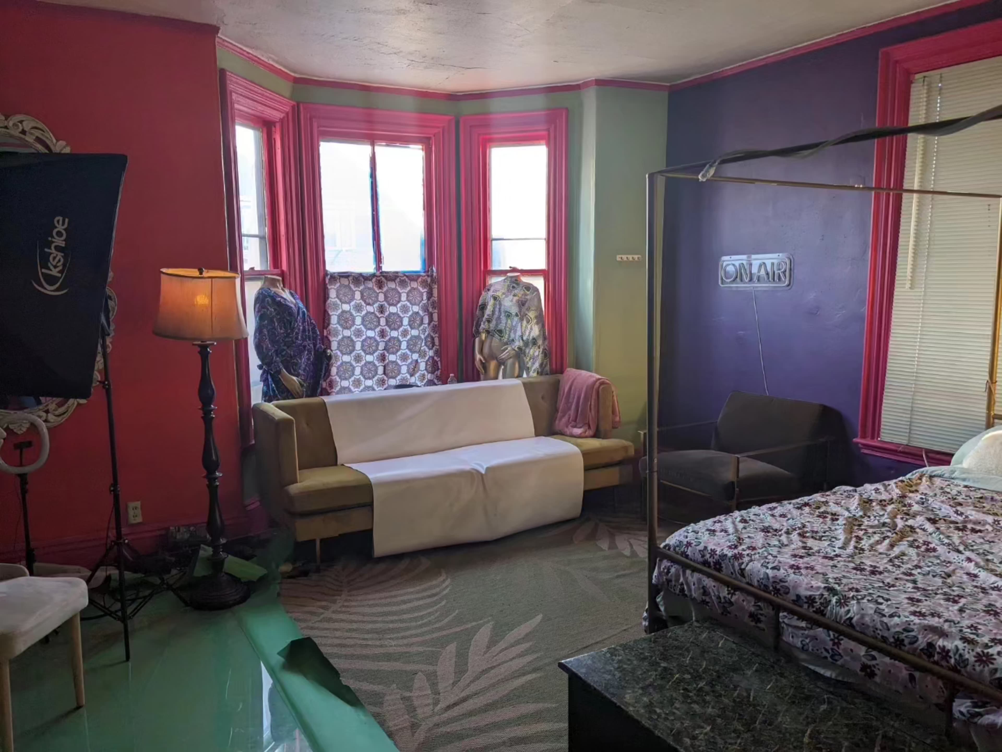 A colorful room featuring a bed with a patterned cover, a sofa partially covered with a white cloth, and a lamp beside a window.