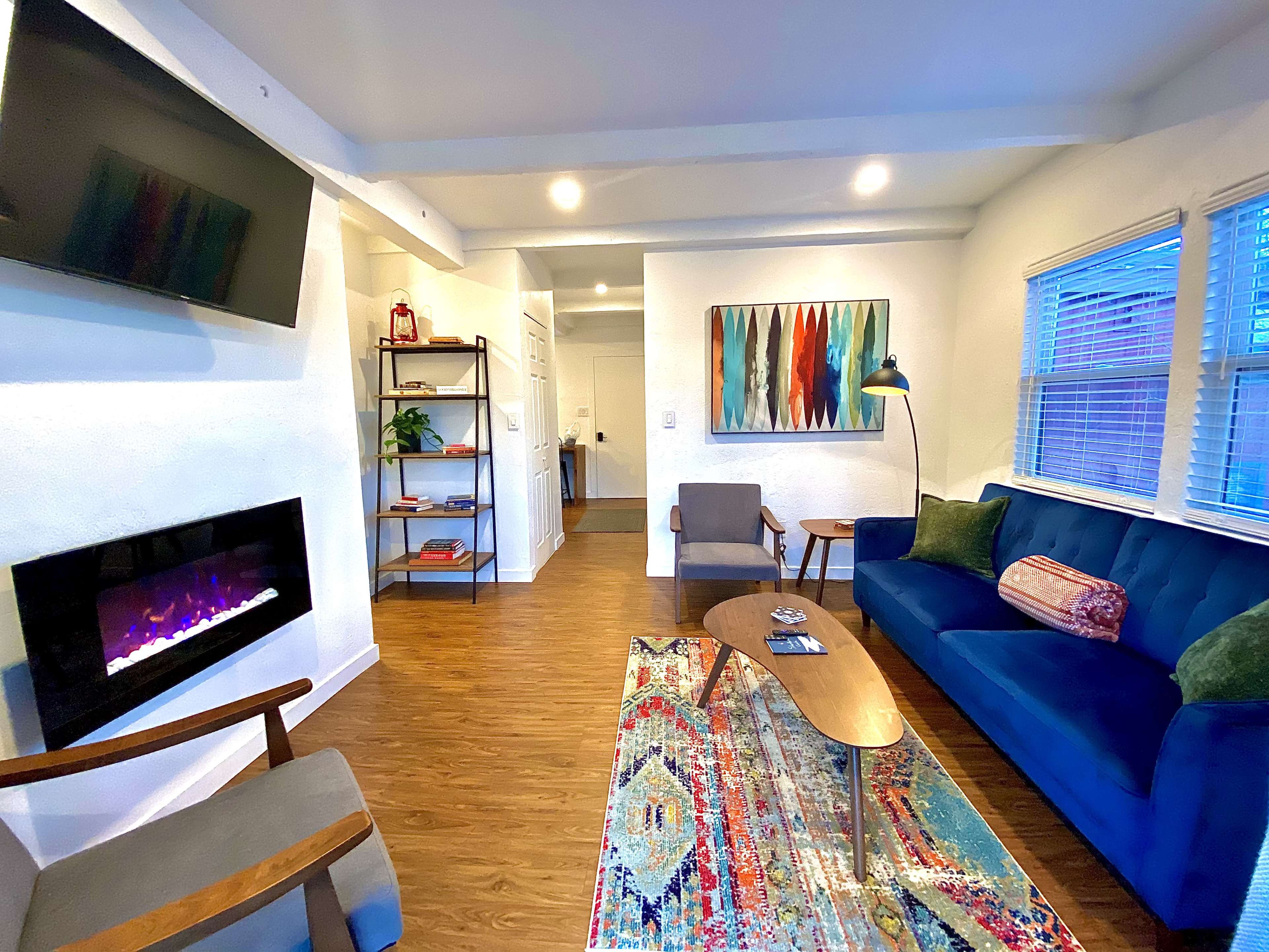 A brightly lit living room with a blue couch, a wooden coffee table, an electric fireplace, and colorful artwork on the wall.