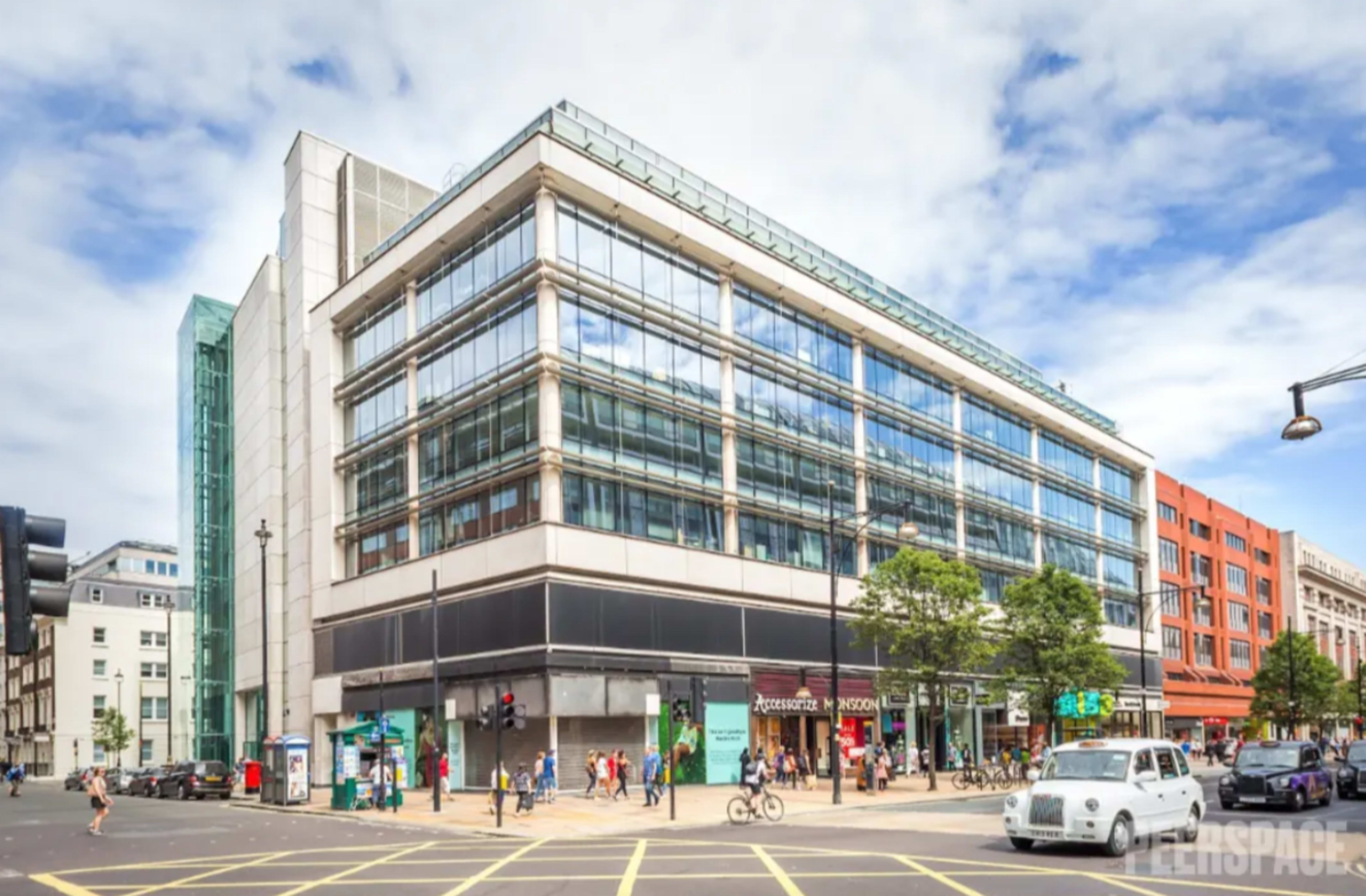 The image shows a modern glass-fronted commercial building at an intersection, with pedestrians and vehicles visible in the foreground.