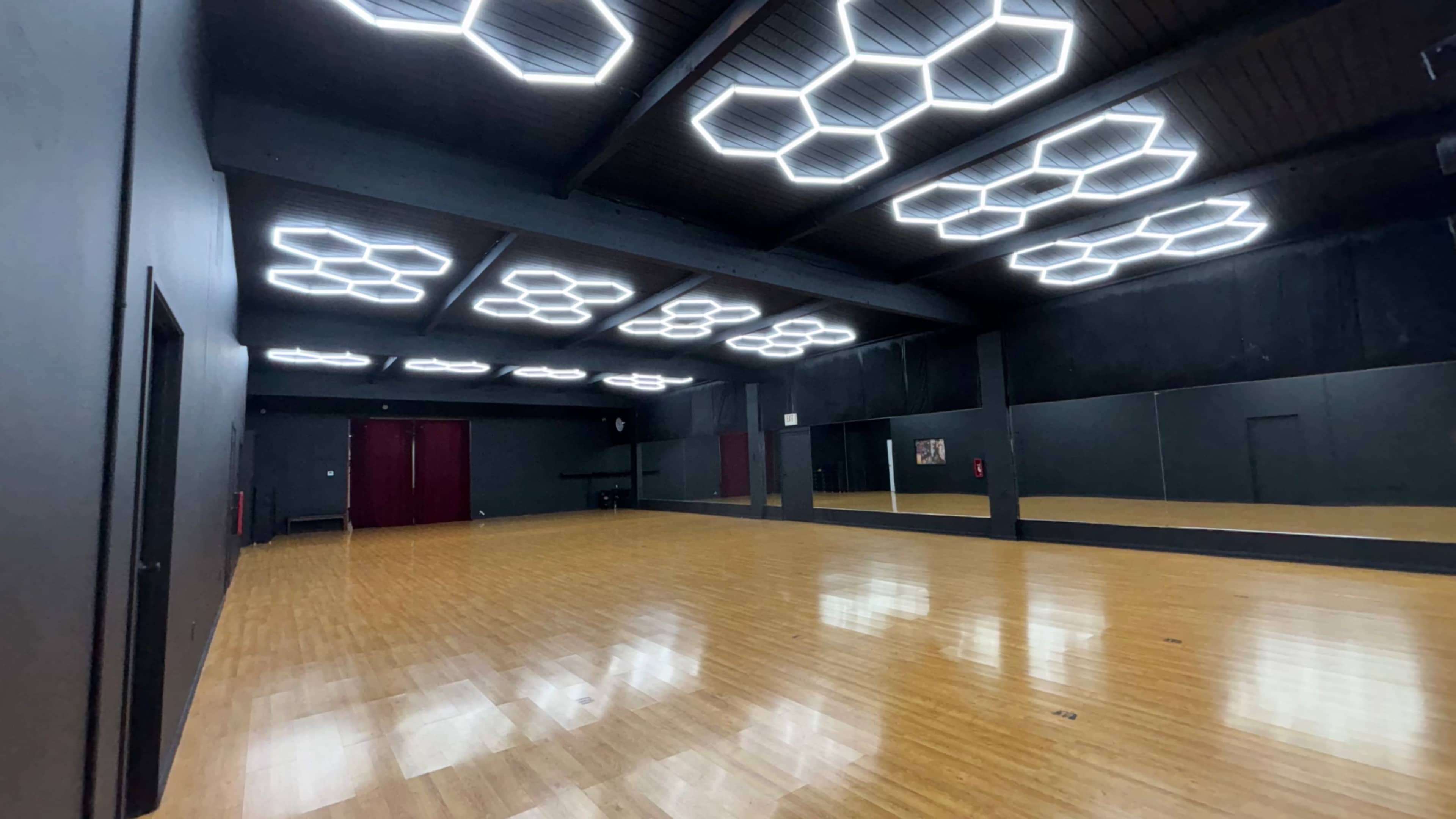 The image shows a spacious dance studio with polished wooden floors, black walls, and modern hexagonal ceiling lights.