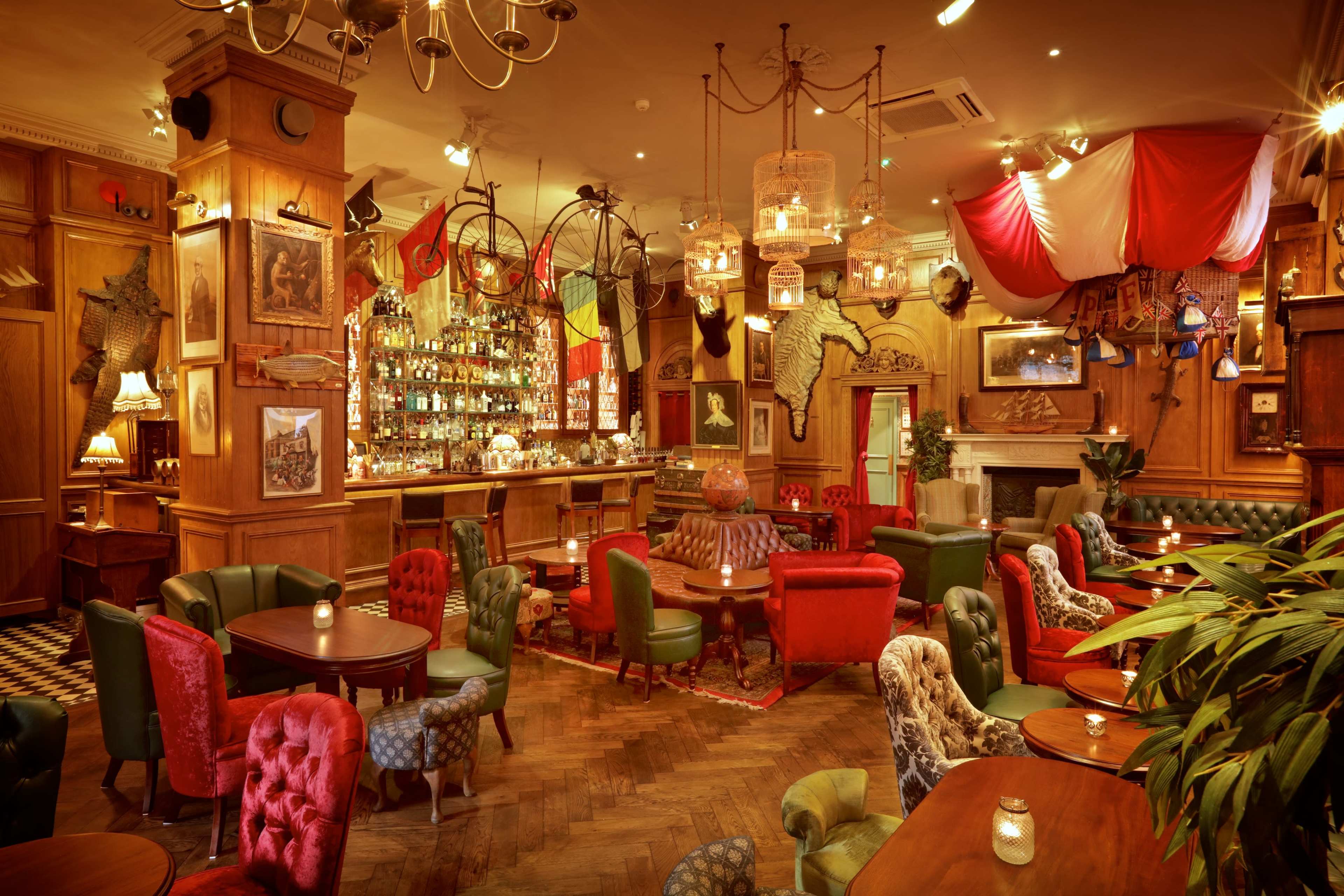 The image shows a cozy bar interior with a mix of colorful upholstered chairs, wooden beams, and a well-stocked bar in the background.