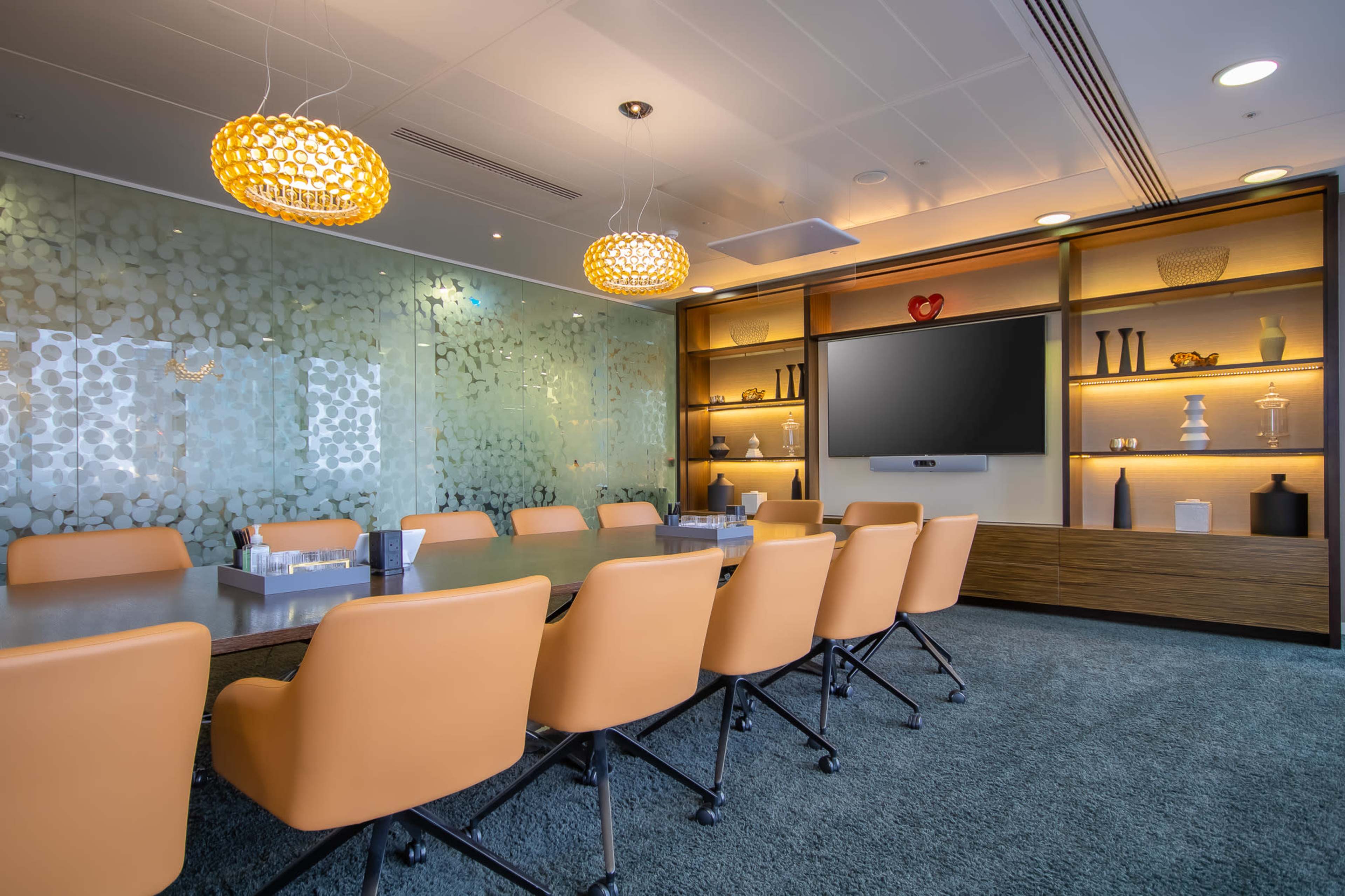 A modern conference room features a long table surrounded by orange chairs, with decorative shelves and a large screen on the wall.