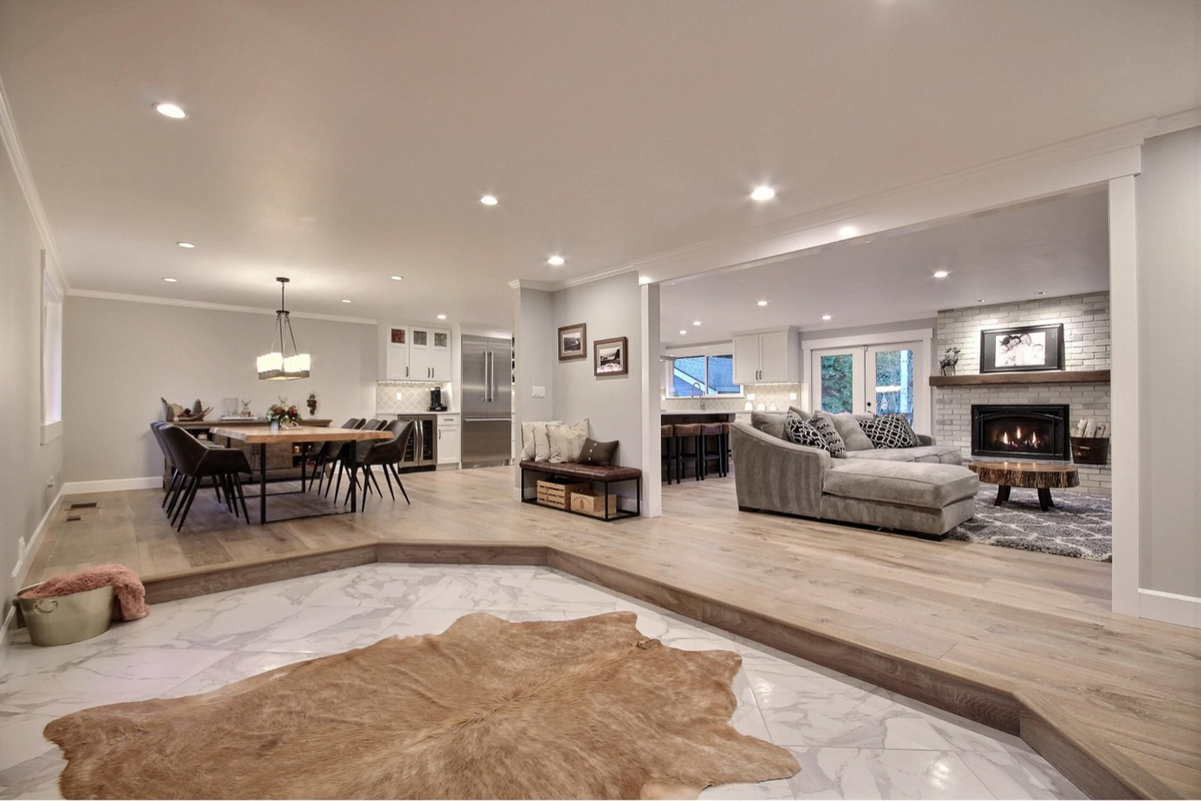 A spacious, open-concept living area features a dining space with a long table, a cozy seating area with a fireplace, and large windows allowing natural light to fill the room.