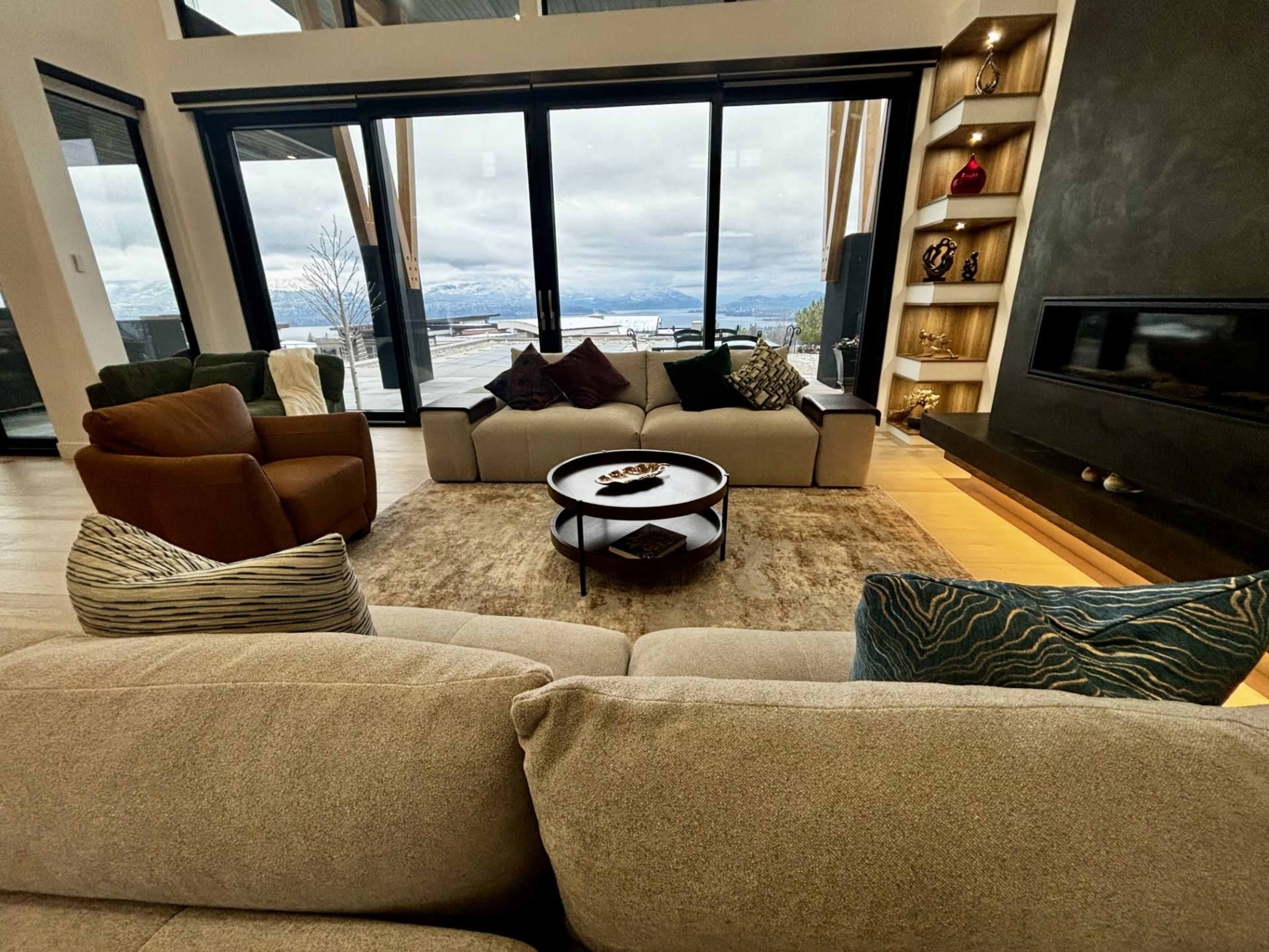 The image shows a modern living room with large windows overlooking a scenic view, featuring a central coffee table surrounded by several couches and armchairs.