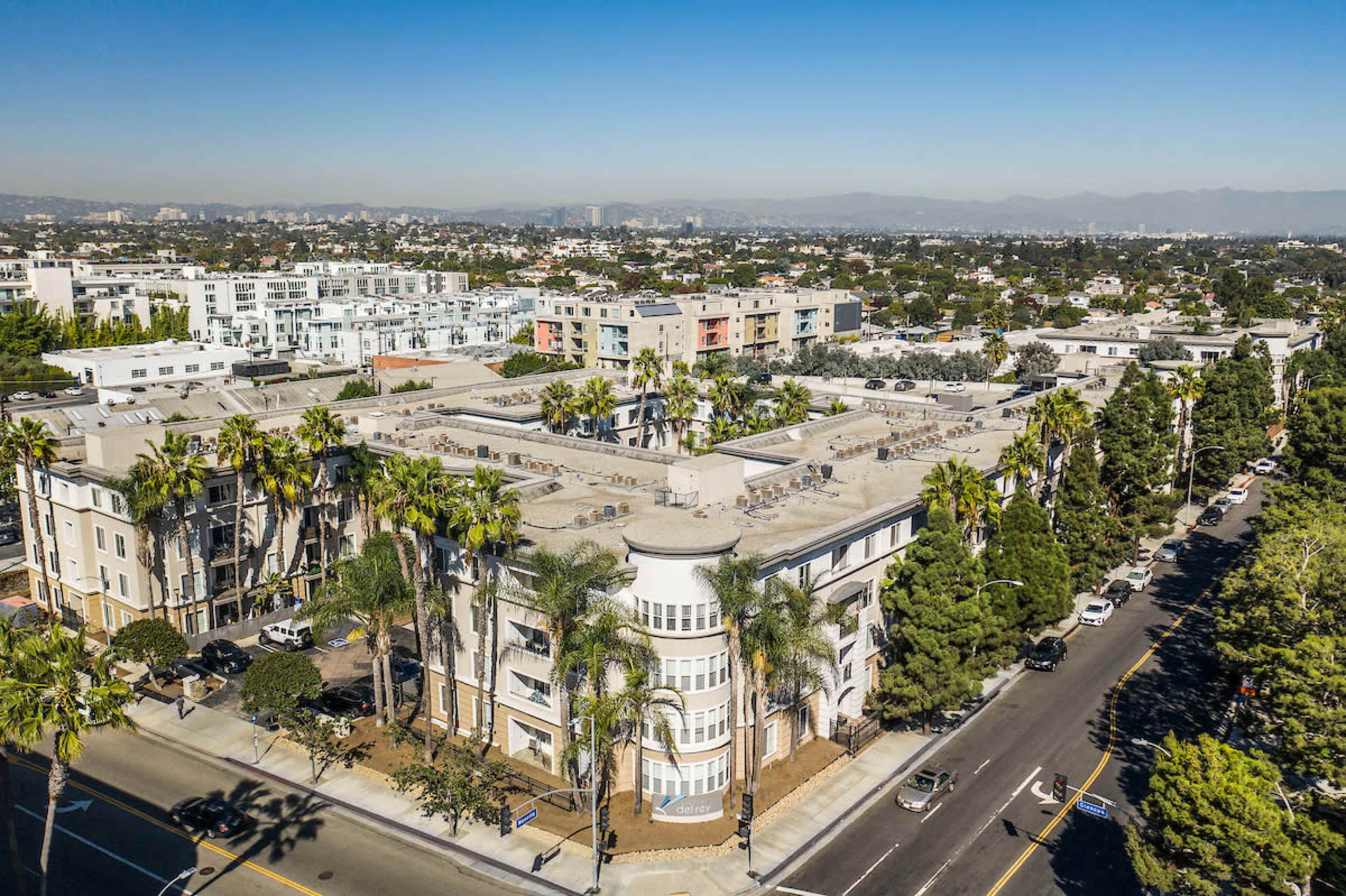 An aerial view shows a cluster of residential buildings surrounded by palm trees and a clear sky.