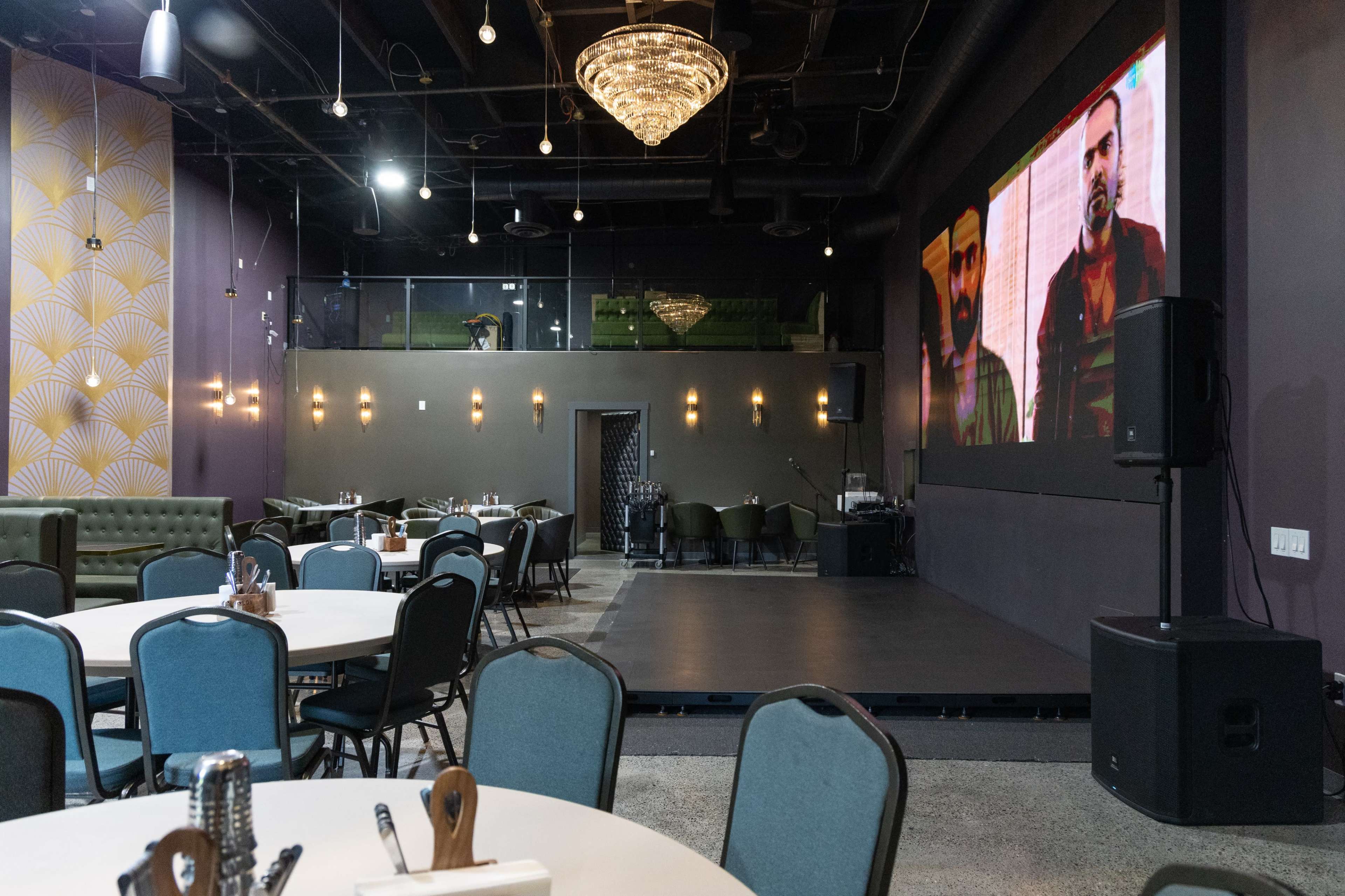 The image shows a modern dining area with circular tables and chairs, a stage featuring a large screen, and decorative wall features.