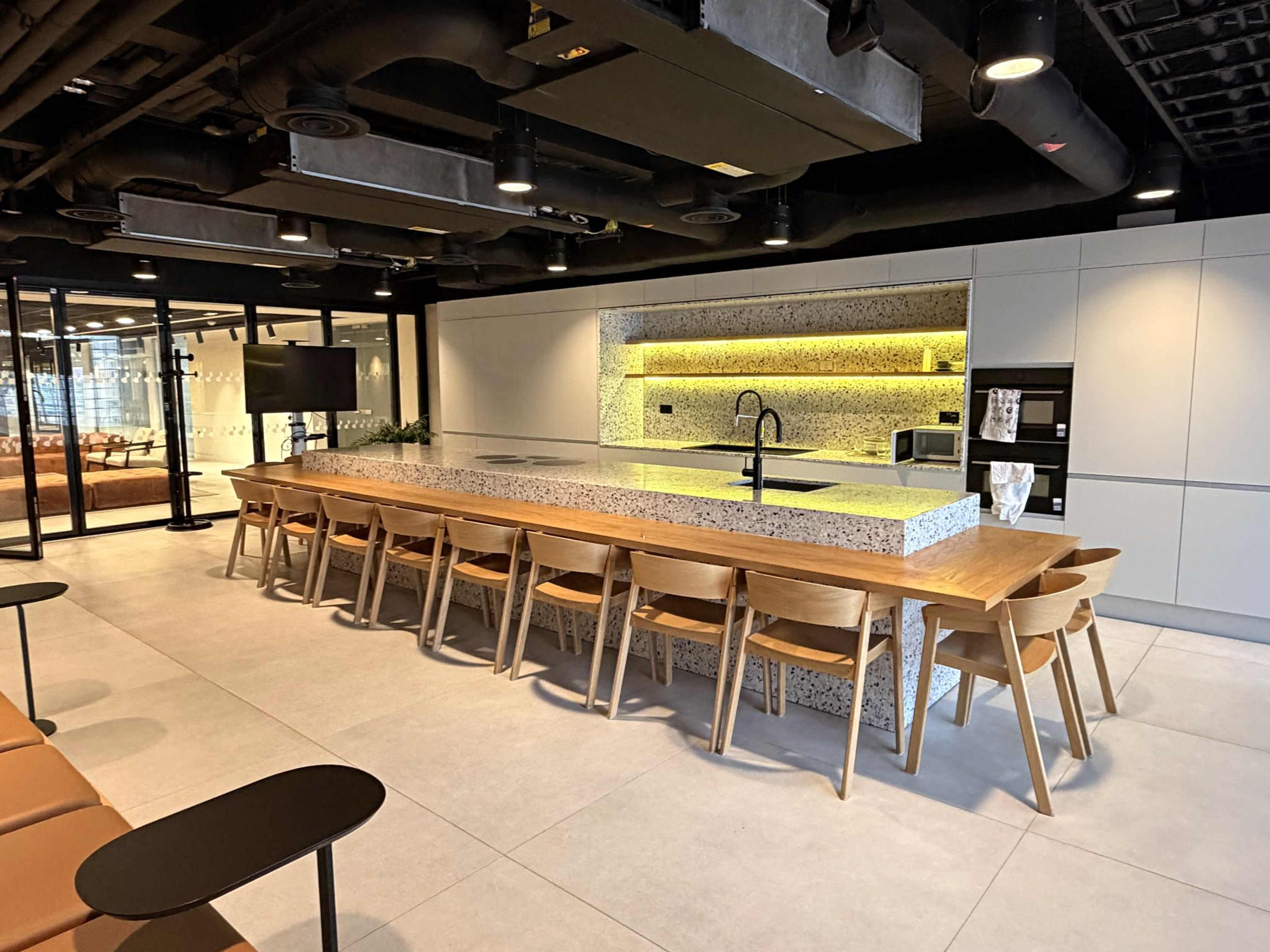The image shows a modern kitchen area with a large stone countertop and wooden seating arranged around it, along with sleek cabinetry and appliances in a spacious, minimalist setting.