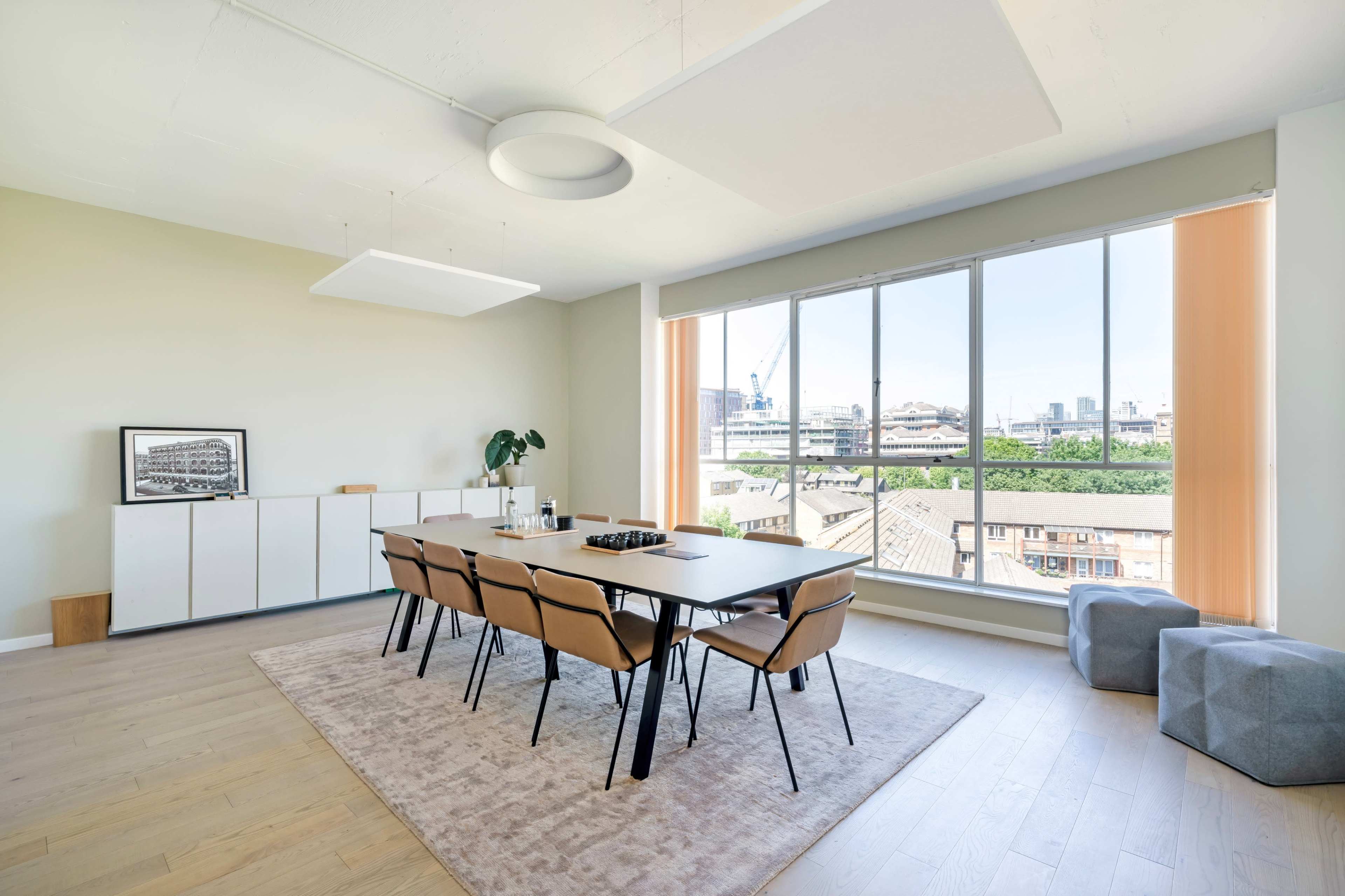 A modern dining area with a long table and chairs, large windows offering a view of the city, and minimalistic furniture.