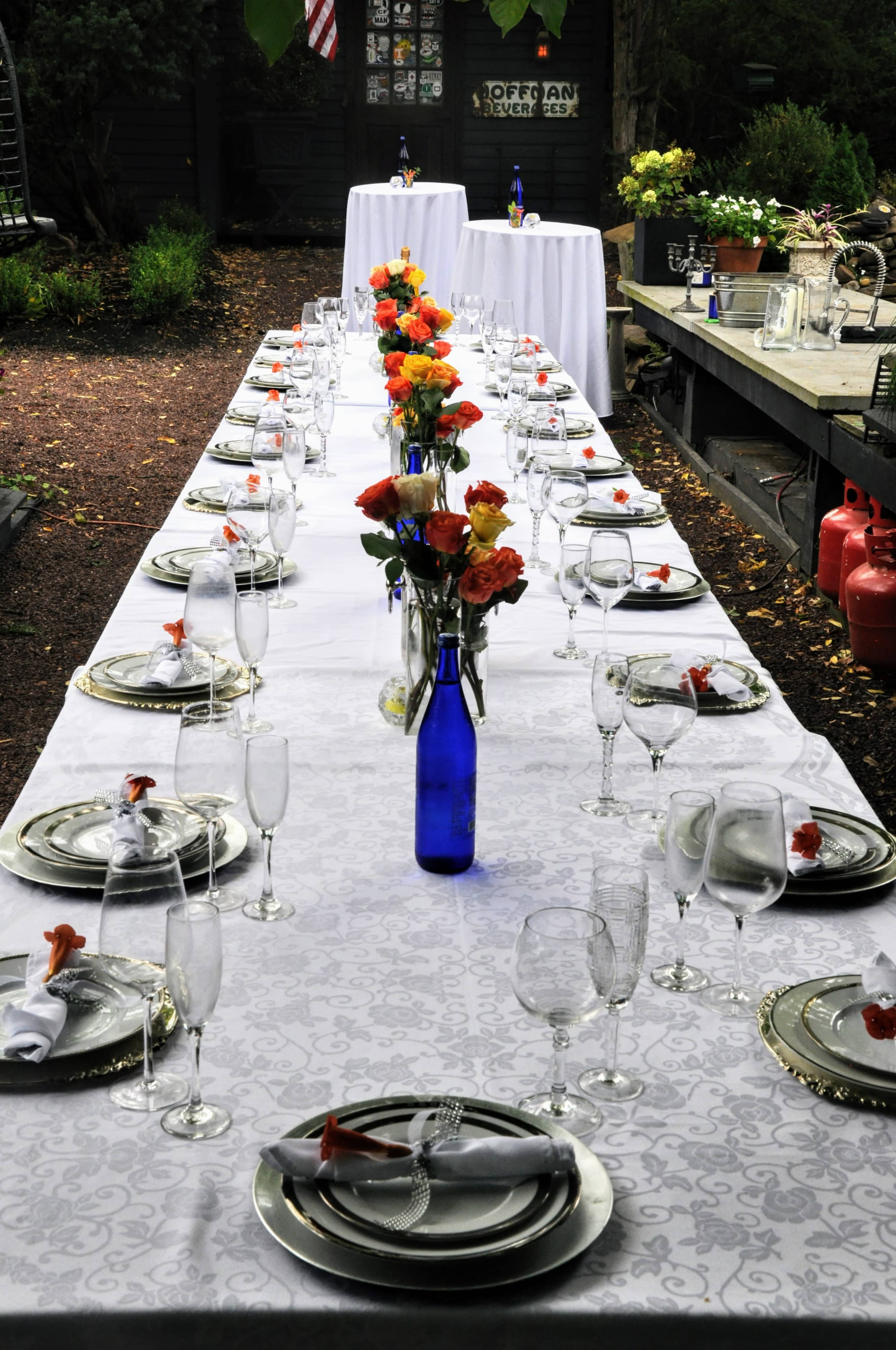 A long, elegantly set outdoor dining table is adorned with floral centerpieces and glassware, ready for a meal.