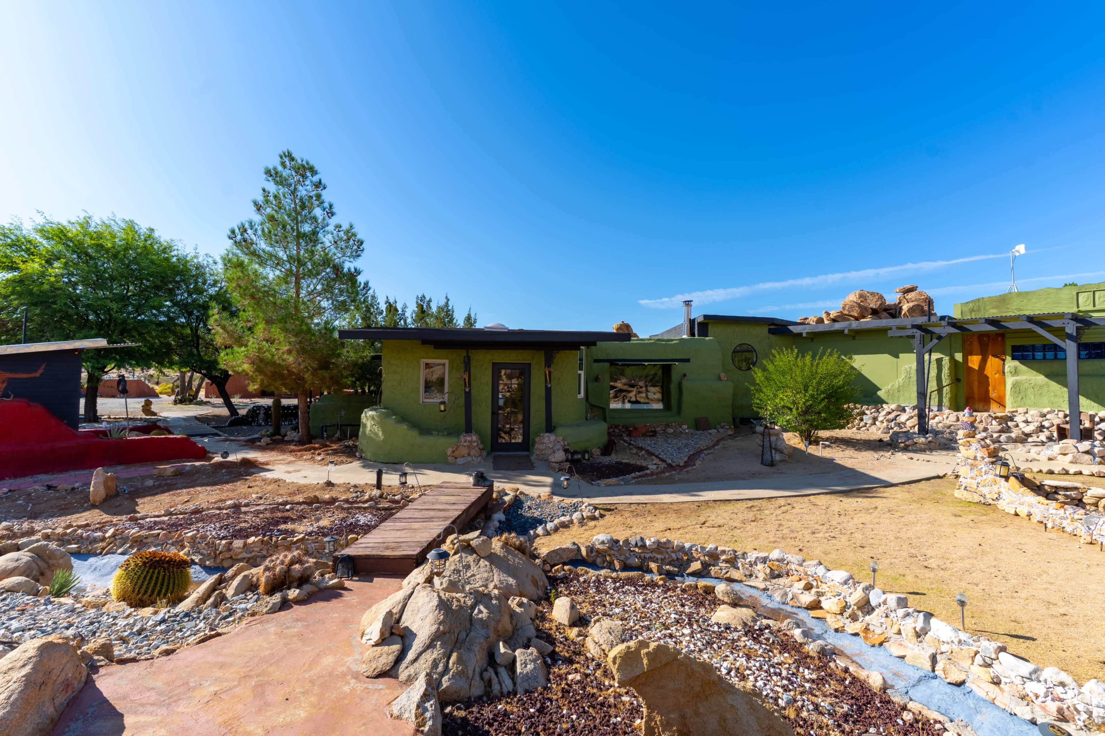 A modern, green-colored house with a stone landscape and a wooden walkway is surrounded by desert vegetation under a clear blue sky.