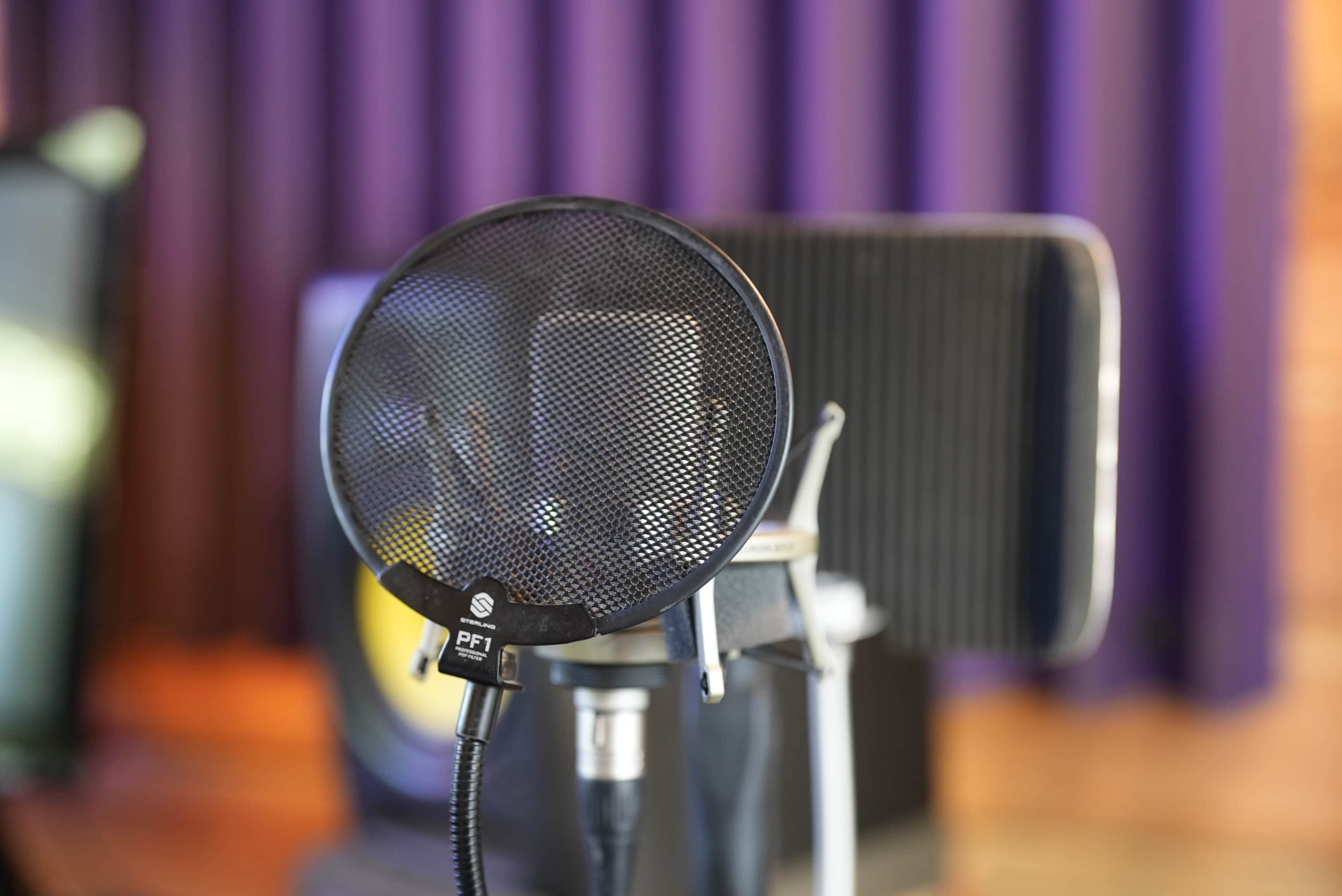 A microphone pop filter is positioned in front of a studio microphone, with blurred speakers and a purple backdrop visible in the background.