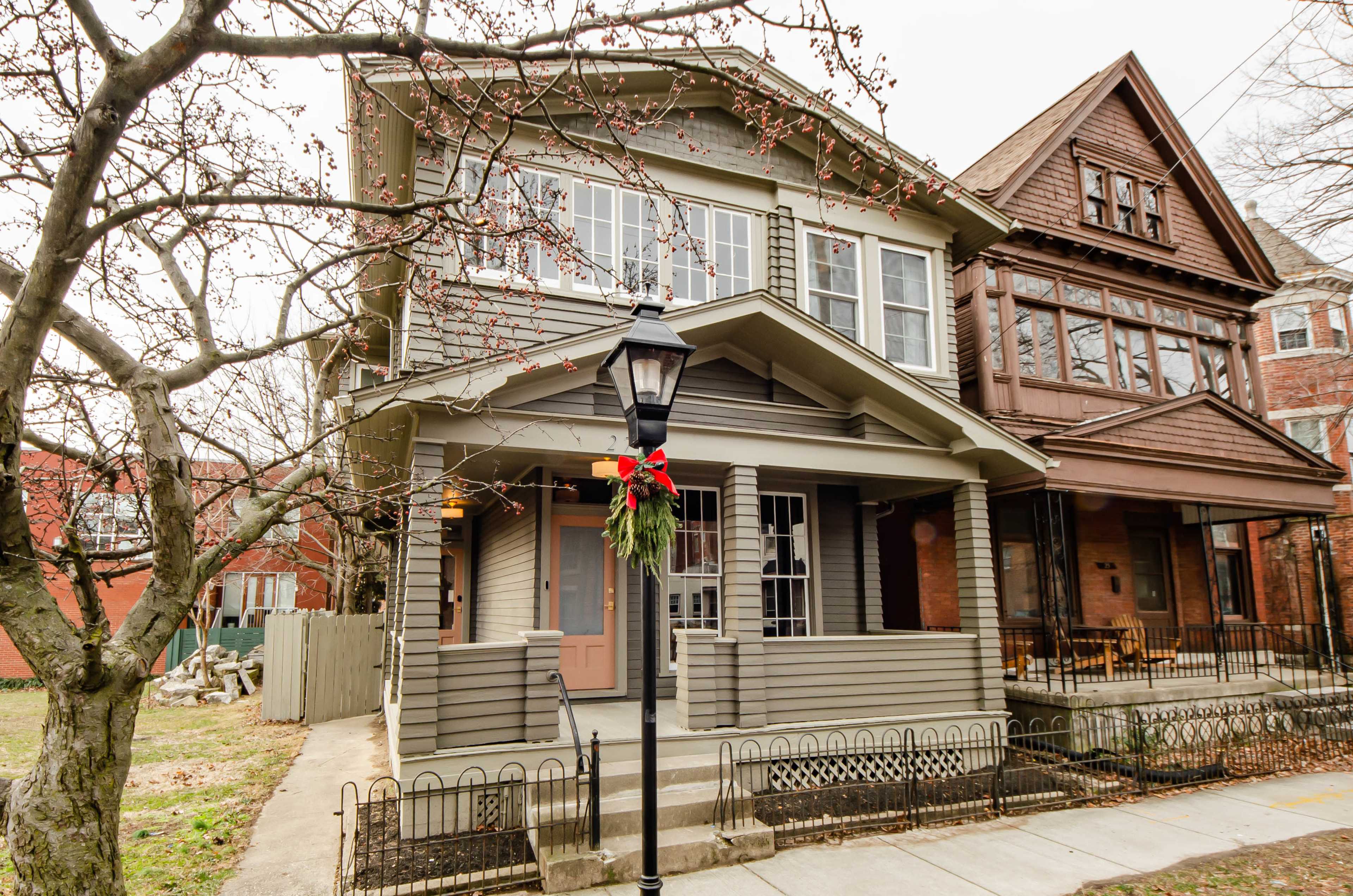 A two-story, gray house with a front porch and decorative holiday wreath on the lamp post is situated next to a larger, brown wooden building.