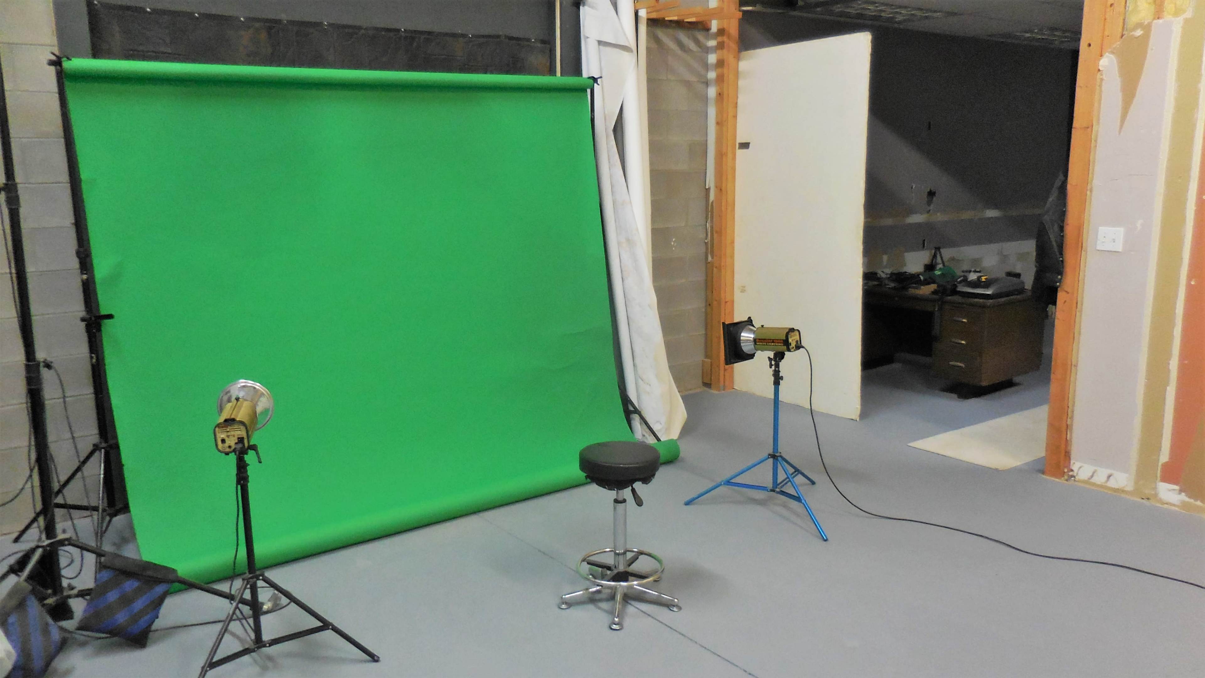 The image shows a studio setup with a green screen, two lights, a stool, and a partially visible workspace in the background.
