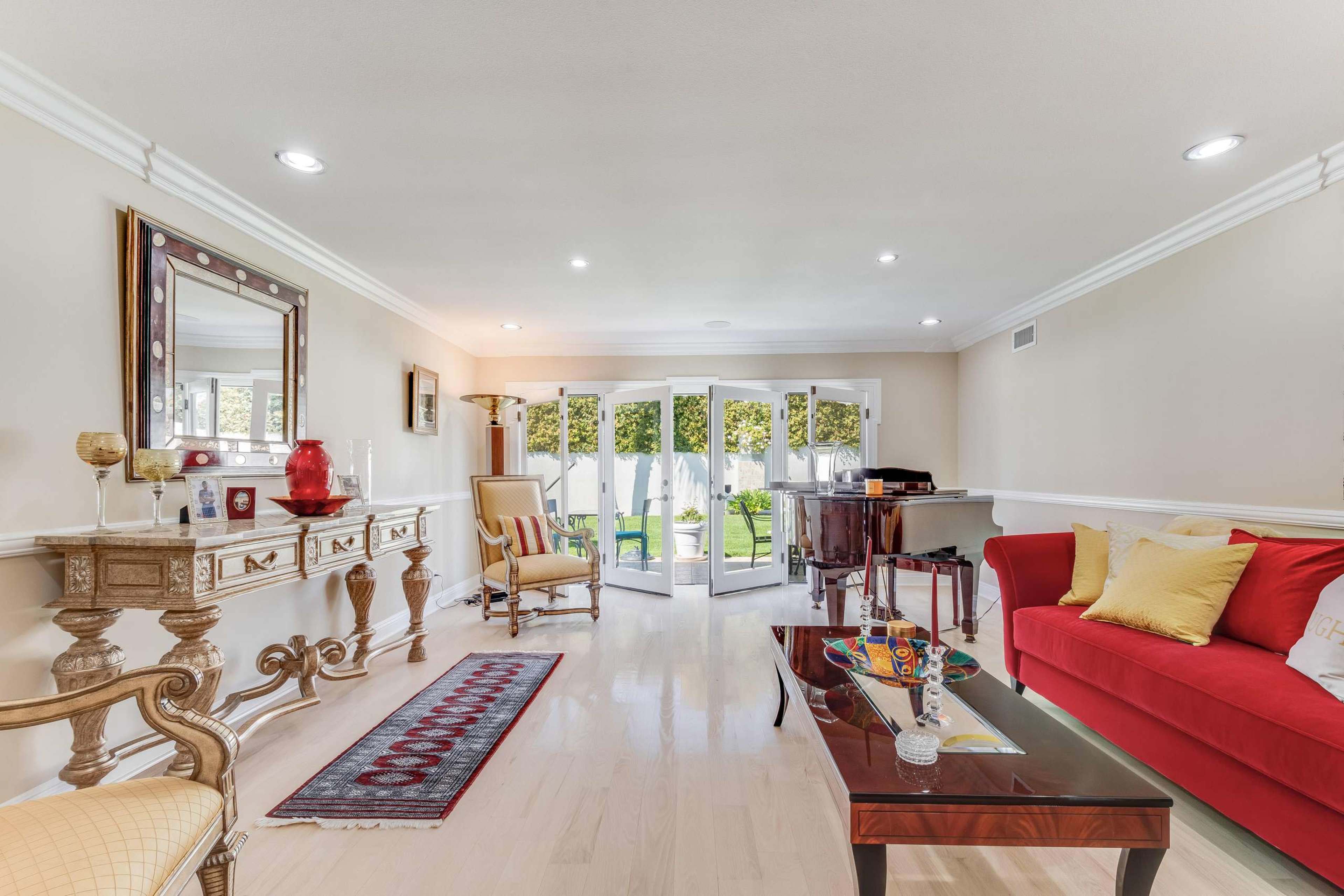 A spacious living room with a red sofa, a wooden piano, a decorative console table, and large glass doors opening to a garden.