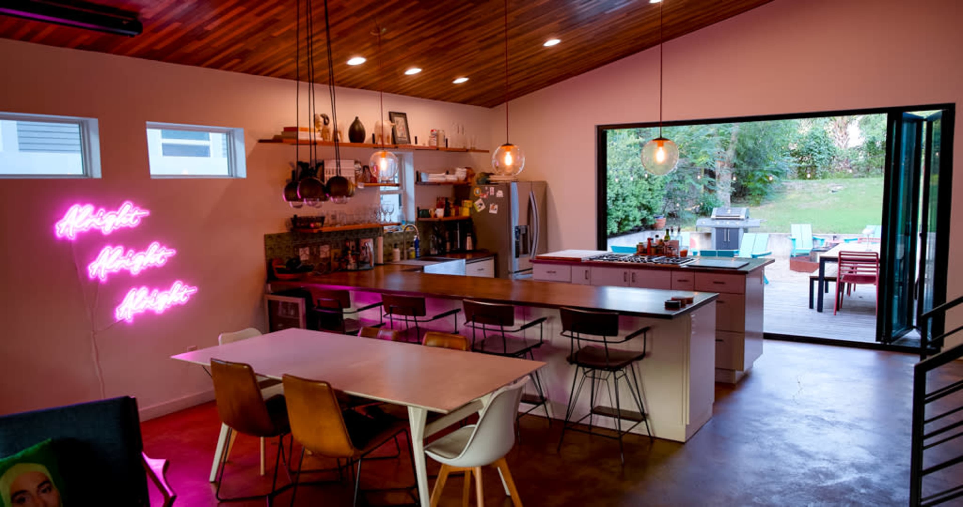 A modern kitchen and dining area features a large central island, wooden ceiling, and an open doorway leading to an outdoor space.