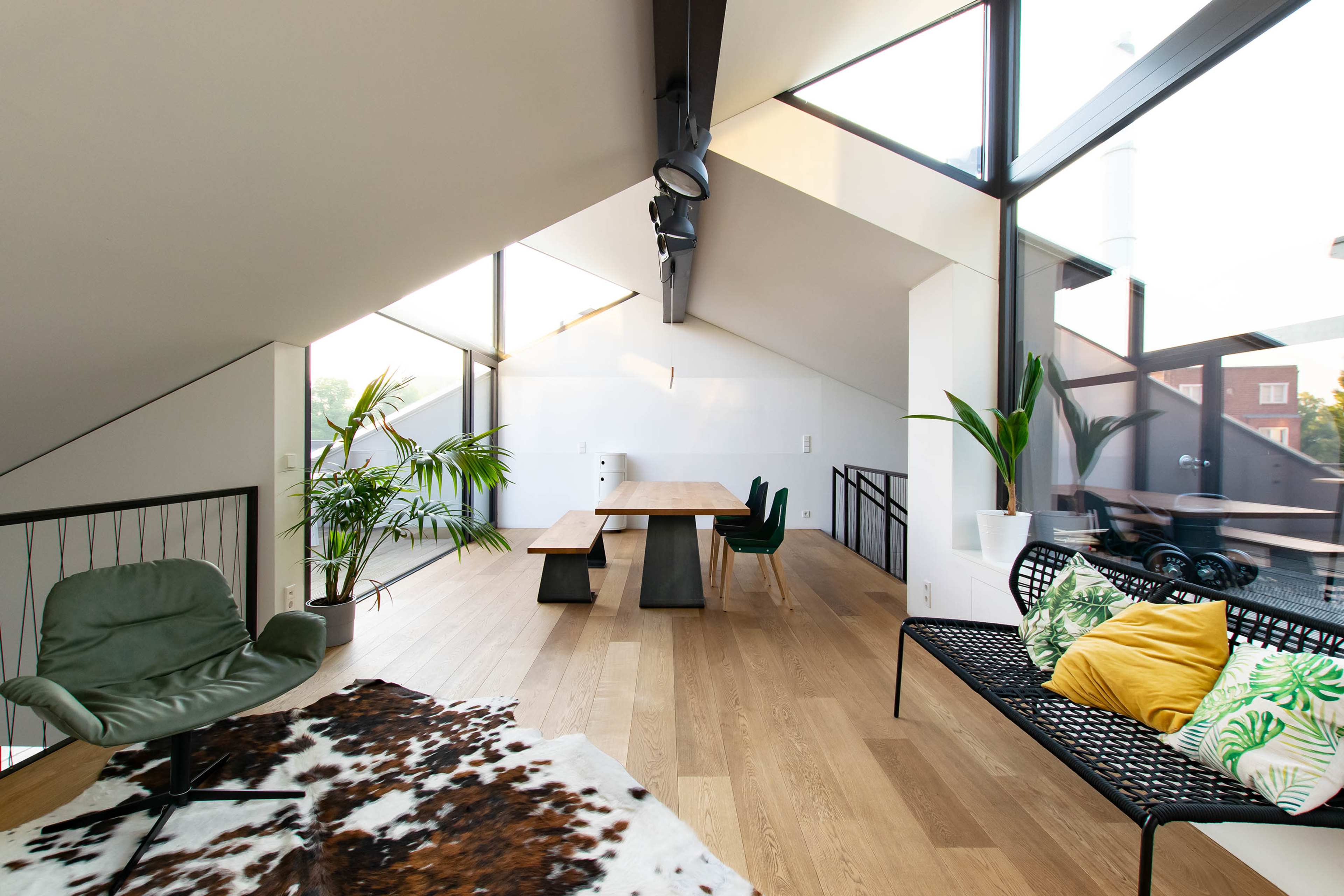 A modern living space features large windows, wooden flooring, a dining table, and a mix of seating options surrounded by indoor plants.