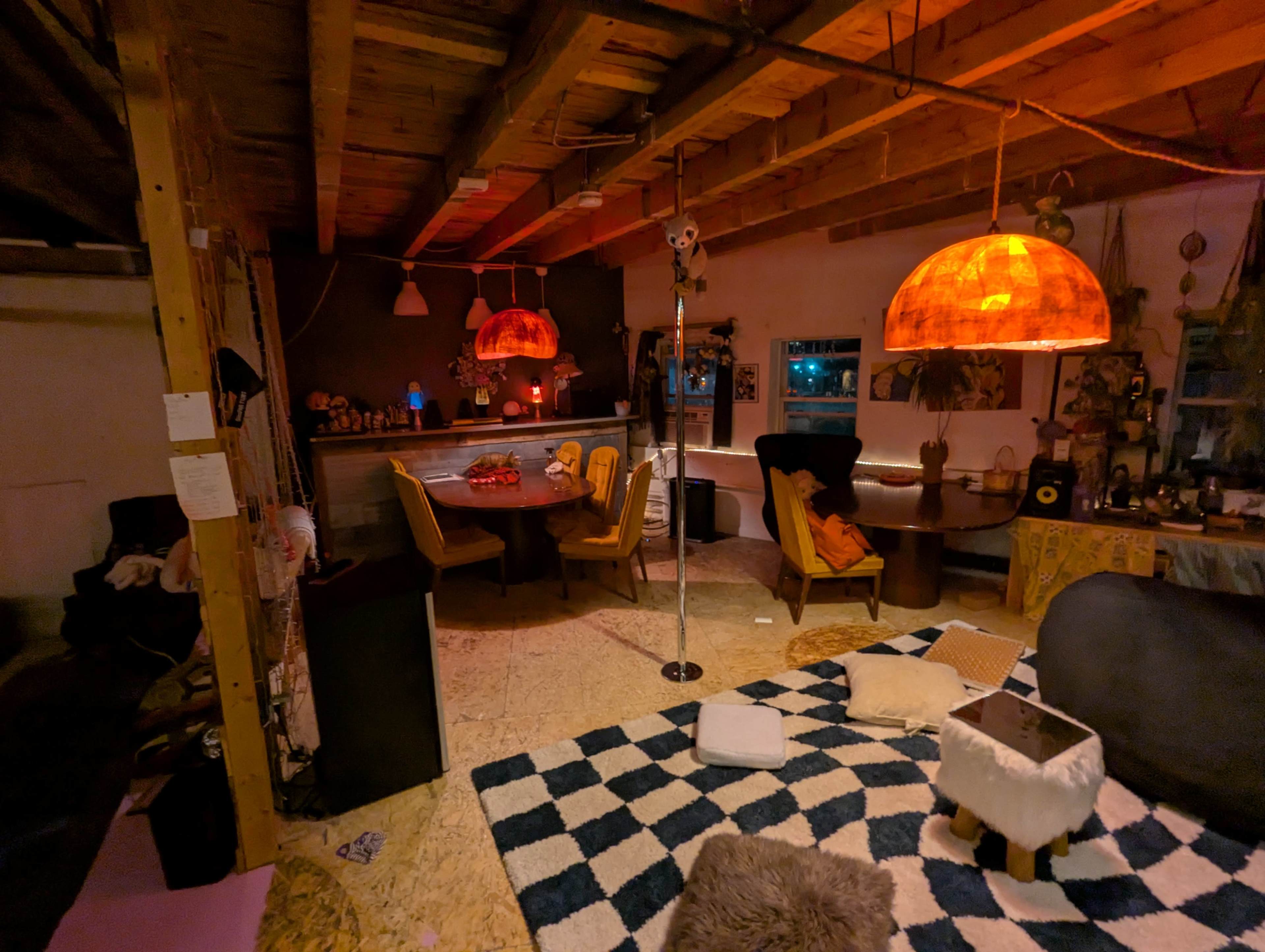 The image shows a cozy living space featuring wooden beams, a dining area with a round table, a lounge area with dark furniture, and two illuminated orange pendant lights hanging from the ceiling.