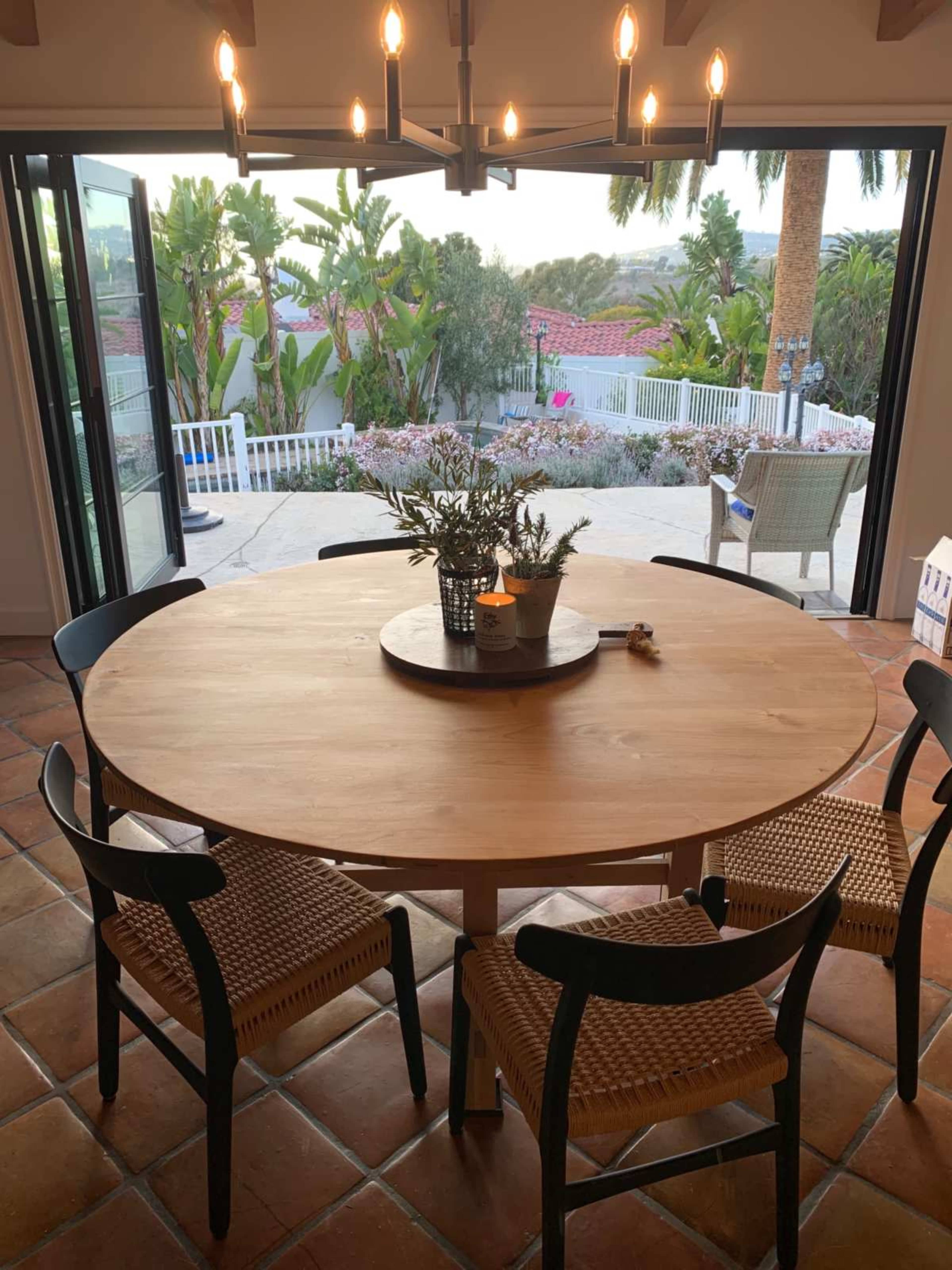 A round wooden dining table with woven chairs is set against a backdrop of an open door, revealing a garden with tropical plants and outdoor seating.