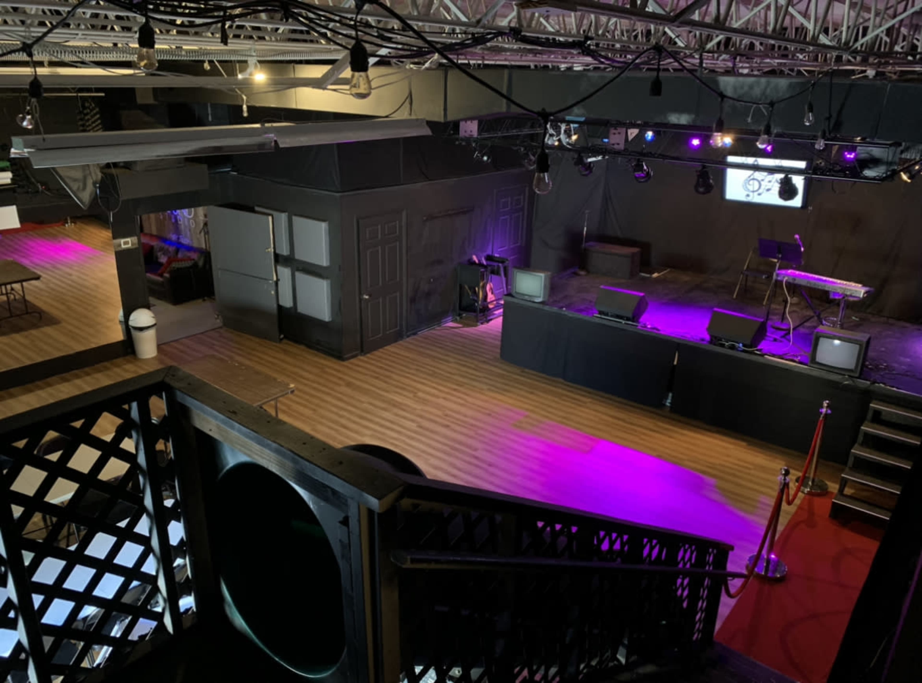 A performance venue features a stage with musical equipment, washed in purple lighting, and a spacious floor lined with tables and chairs.