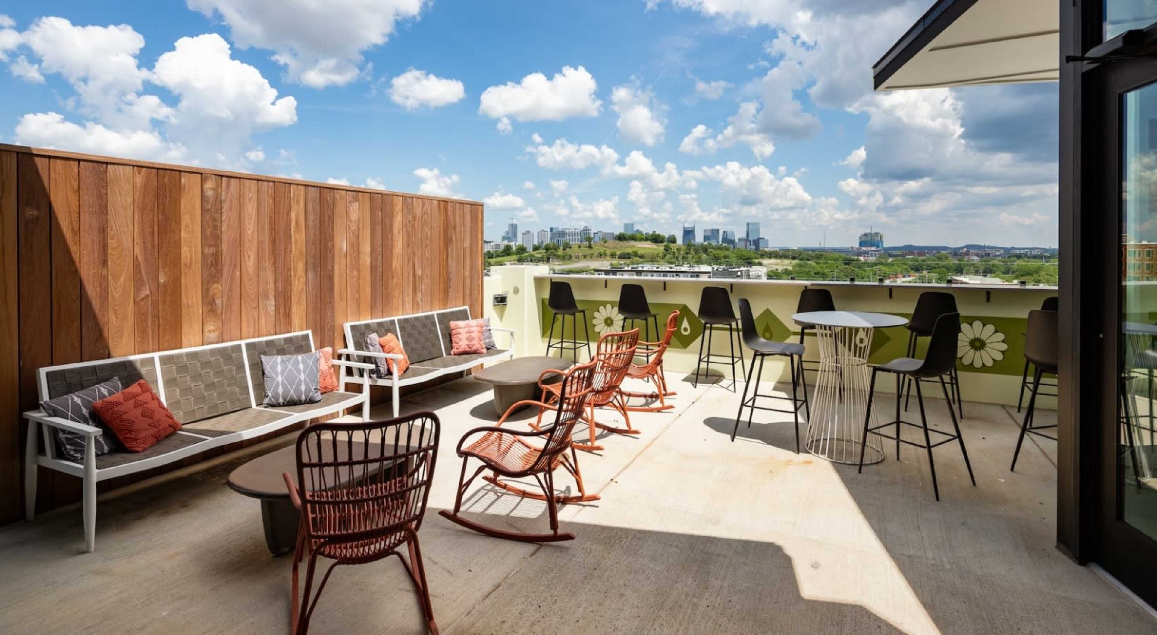 The image shows a rooftop terrace with various seating arrangements overlooking a city skyline under a blue sky with fluffy white clouds.
