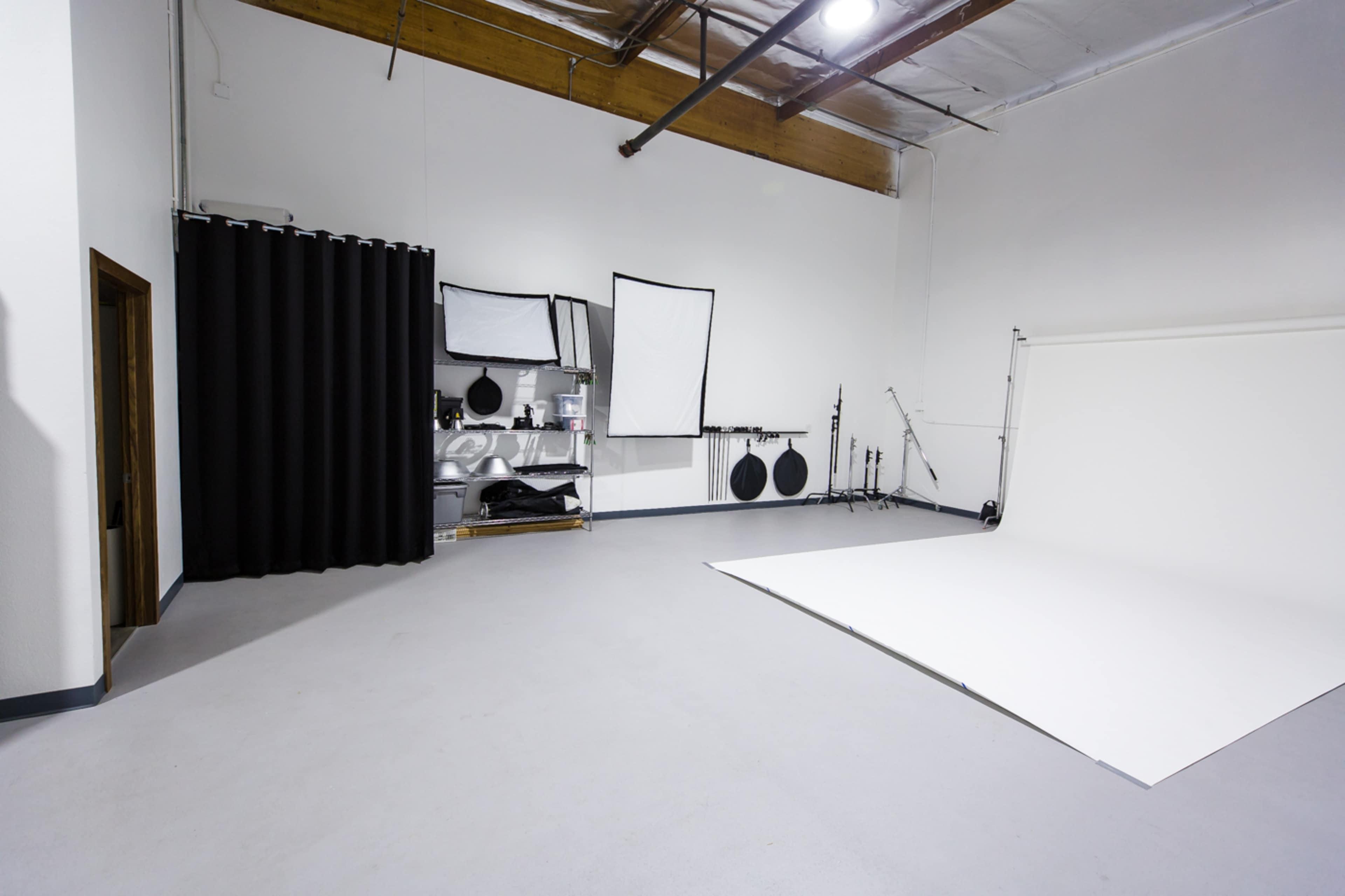 A spacious studio with white walls, a white backdrop, black curtains, and various photography equipment on shelves.