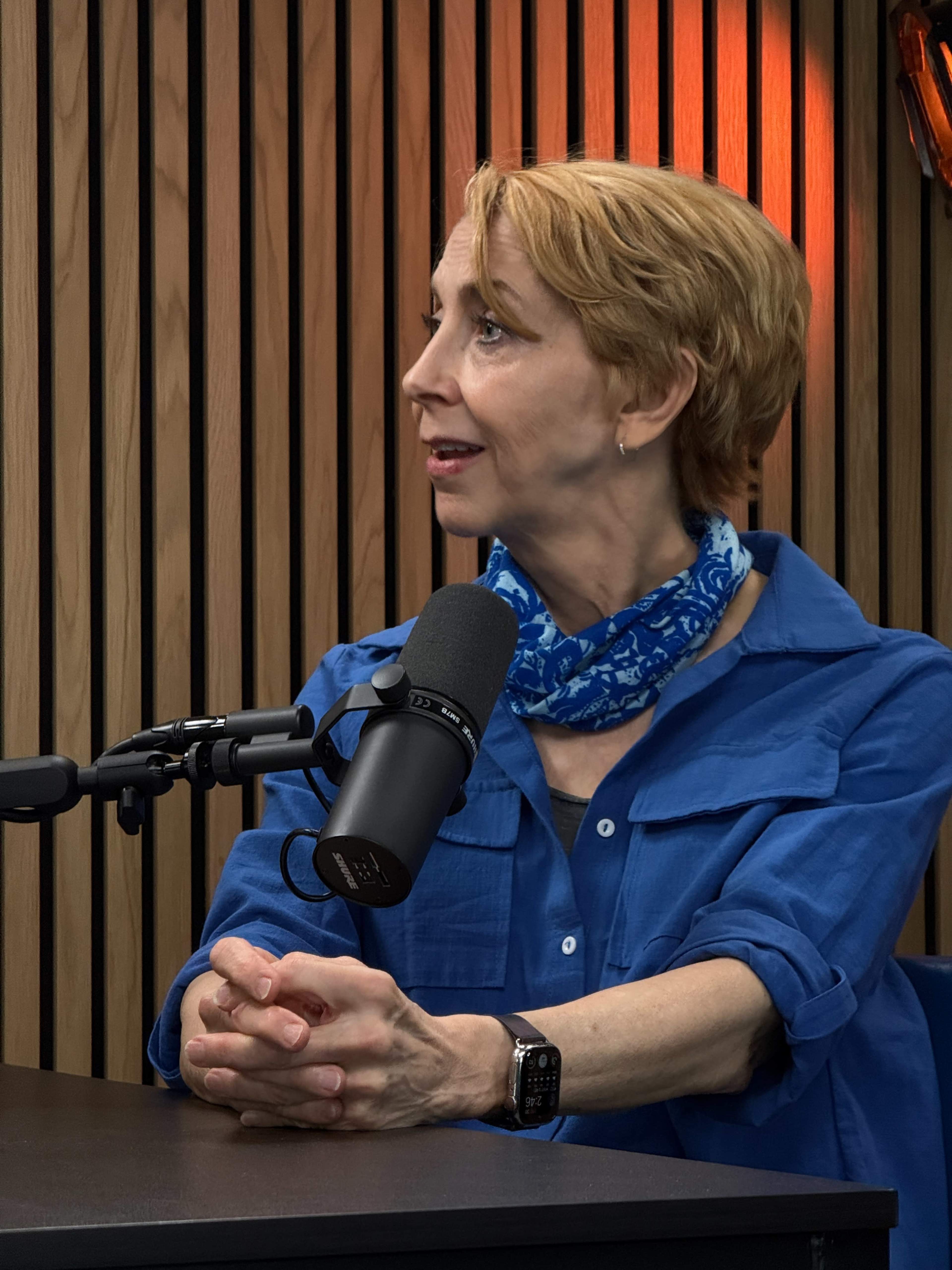 A woman with short blonde hair sits at a podcast table, speaking into a microphone while wearing a blue shirt and a patterned scarf.