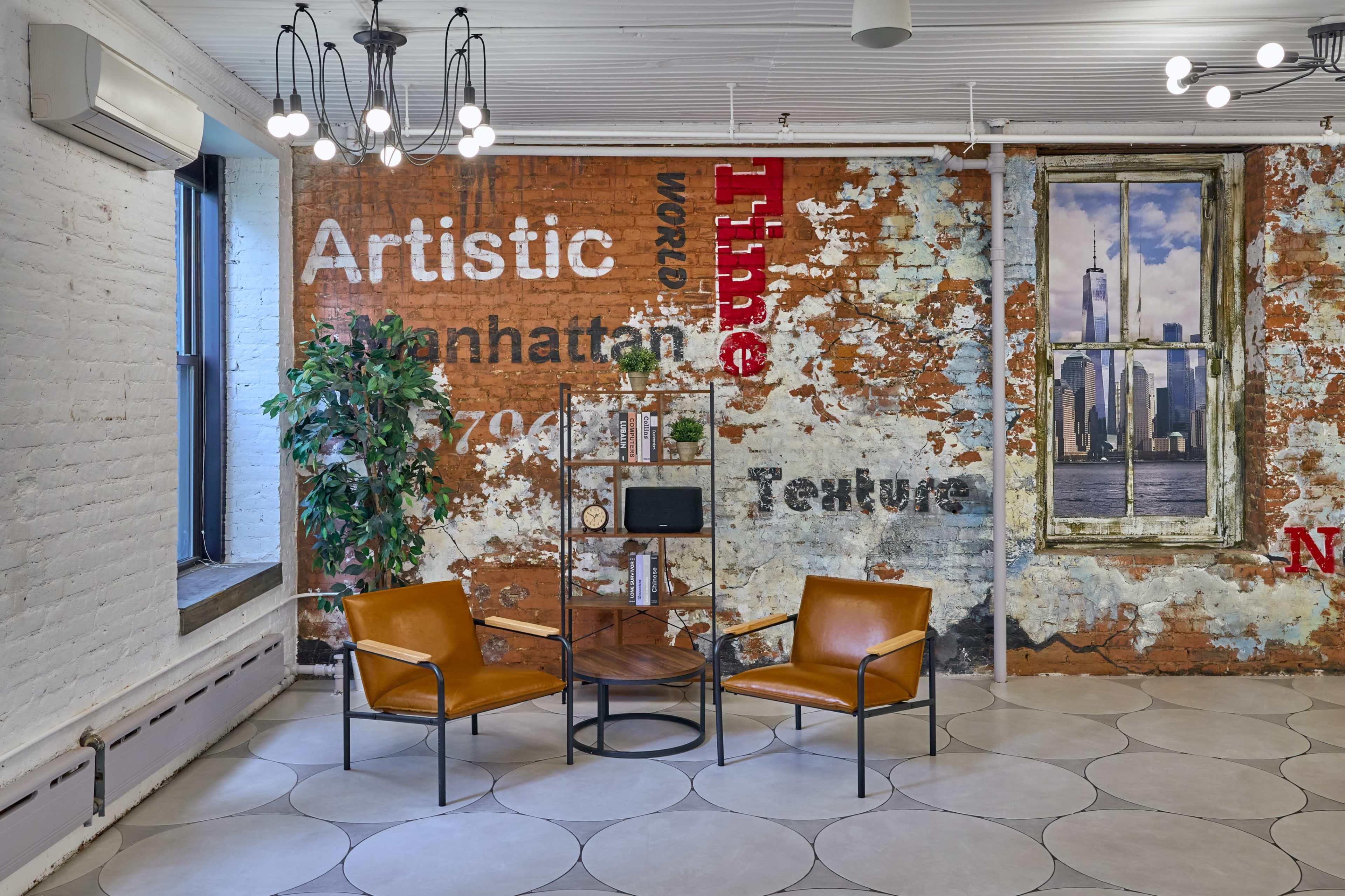 The image shows a contemporary indoor space featuring two leather chairs, a round table, a decorative plant, and a wall with a textured design and window view.
