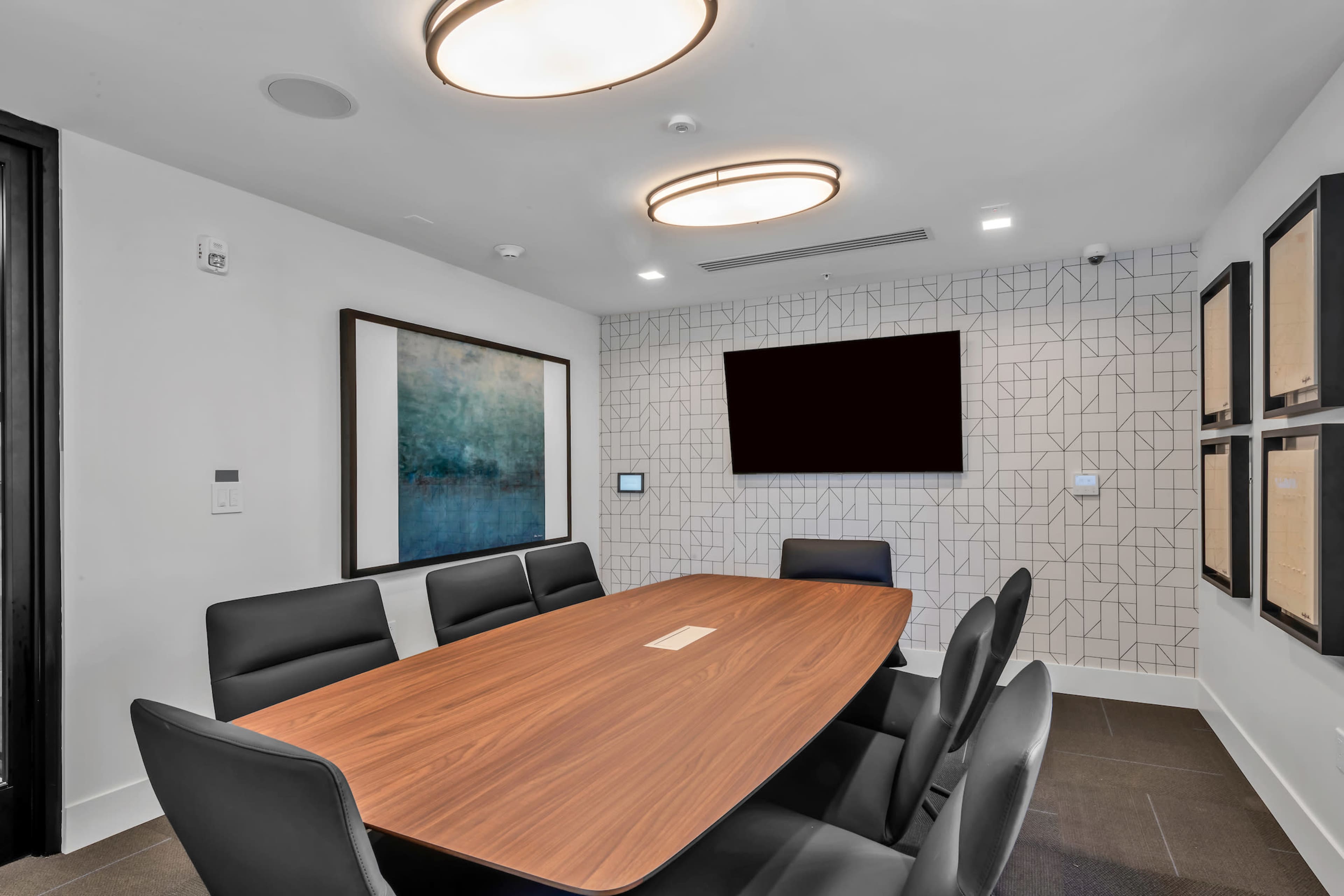 A modern conference room features a large wooden table surrounded by black chairs, with a wall-mounted screen and geometric patterned decor.