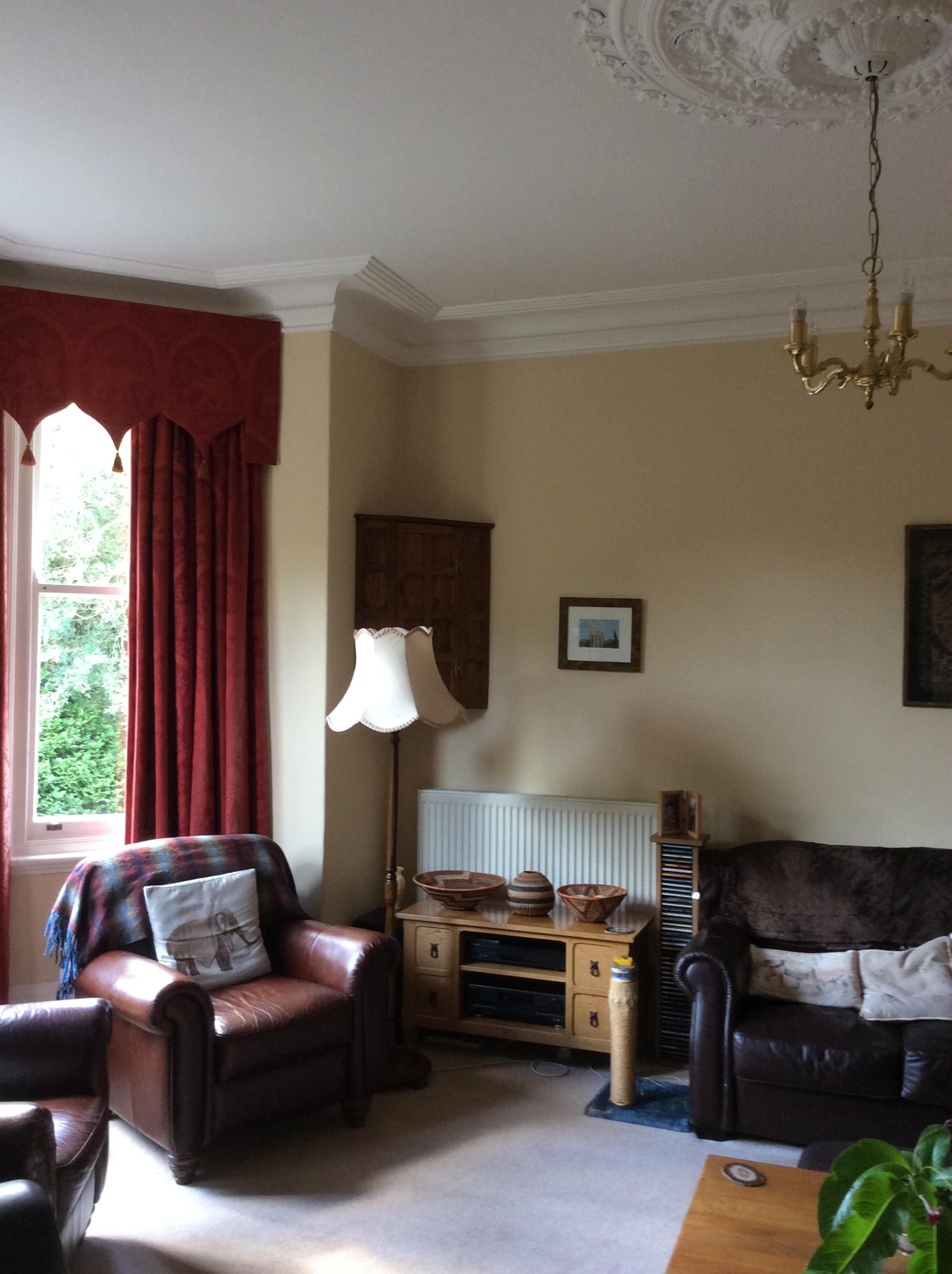 A living room featuring two leather armchairs, a lamp, a radiator, and a wooden cabinet, with red curtains and cream walls.