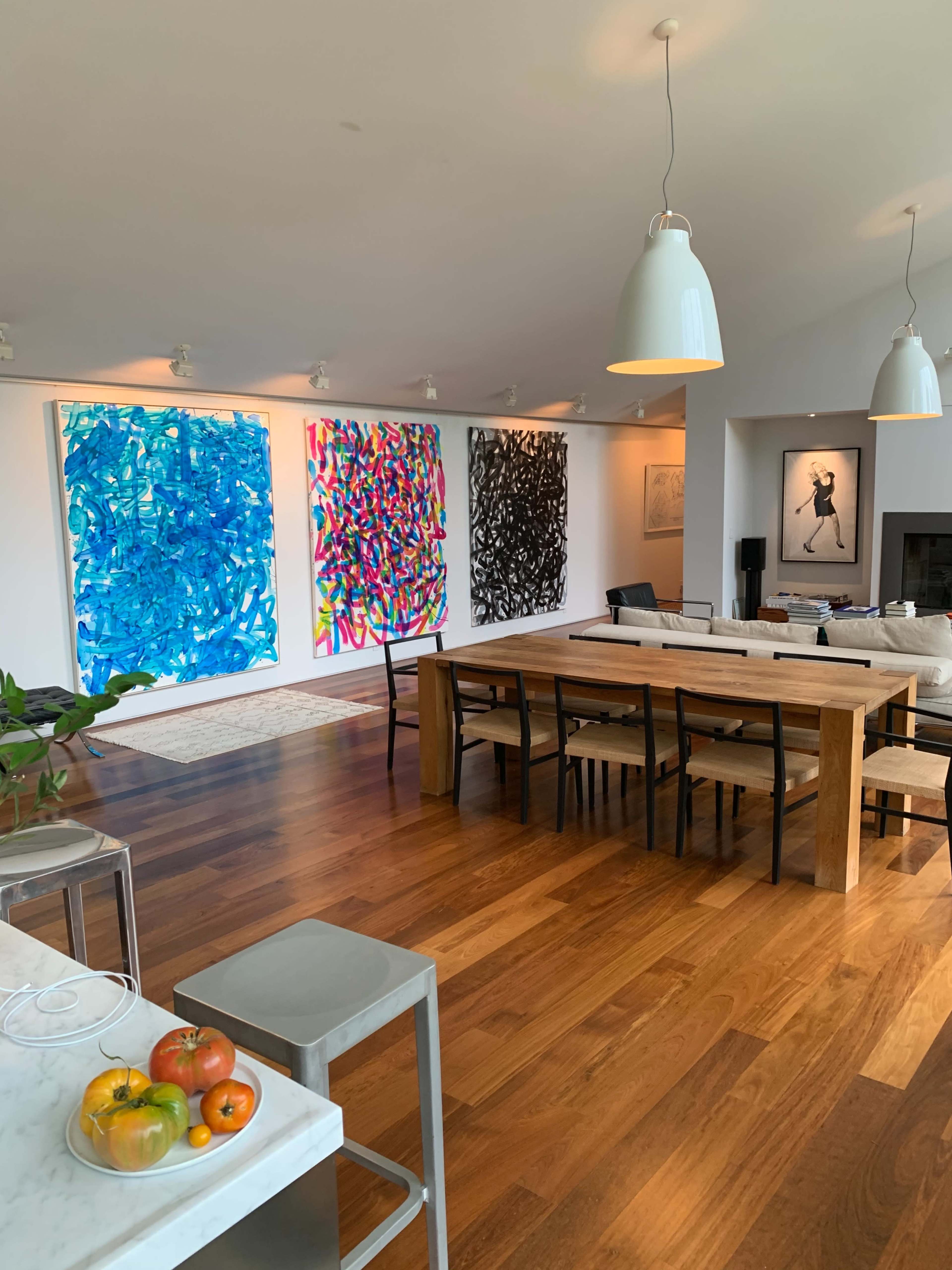 A spacious dining area with a long wooden table, surrounded by chairs, and three large abstract paintings on the wall, alongside a living area that features a fireplace and a small table with fruit.