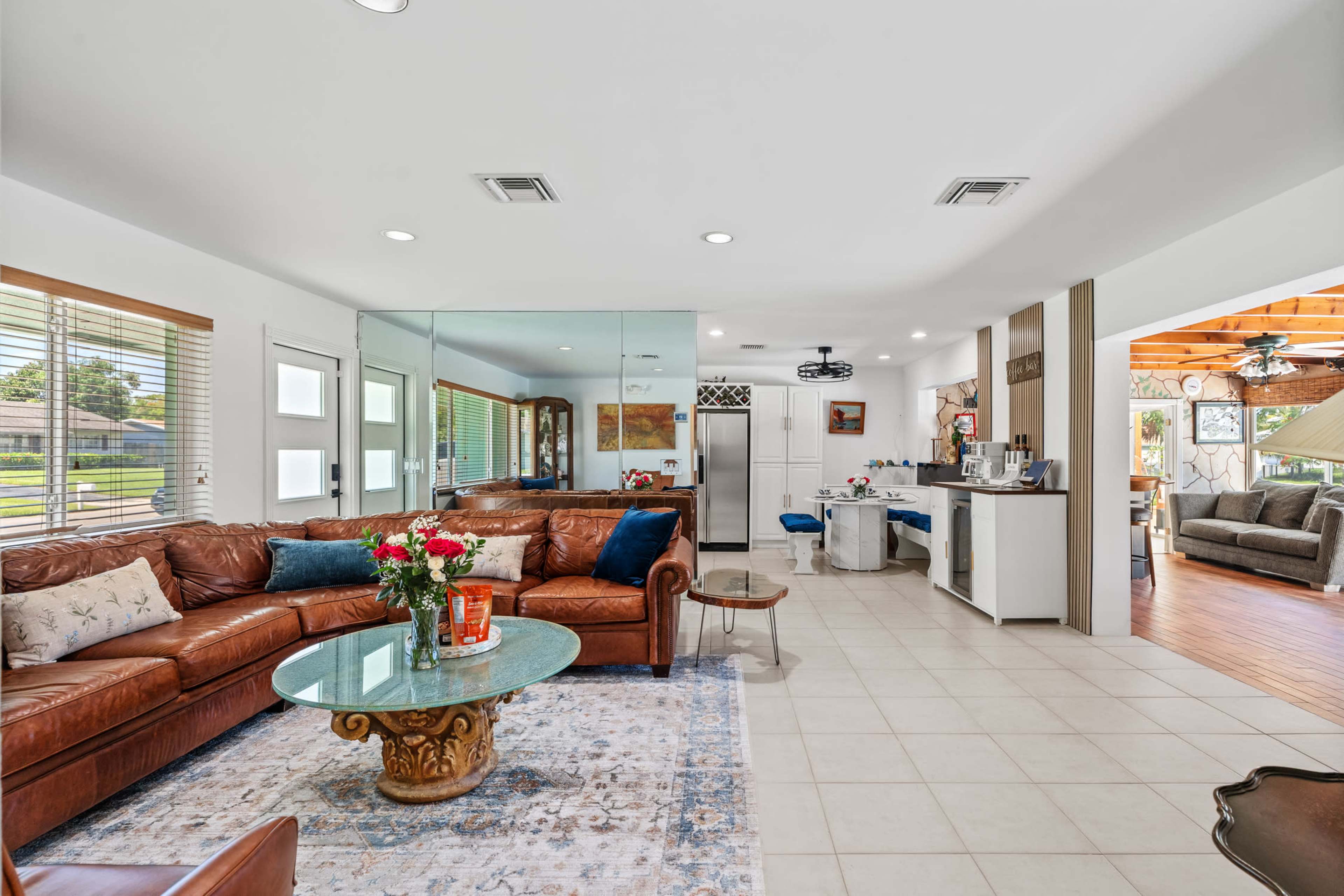 The image shows a bright, open living space featuring a leather sectional sofa, a glass coffee table with a floral arrangement, and a kitchen area in the background.