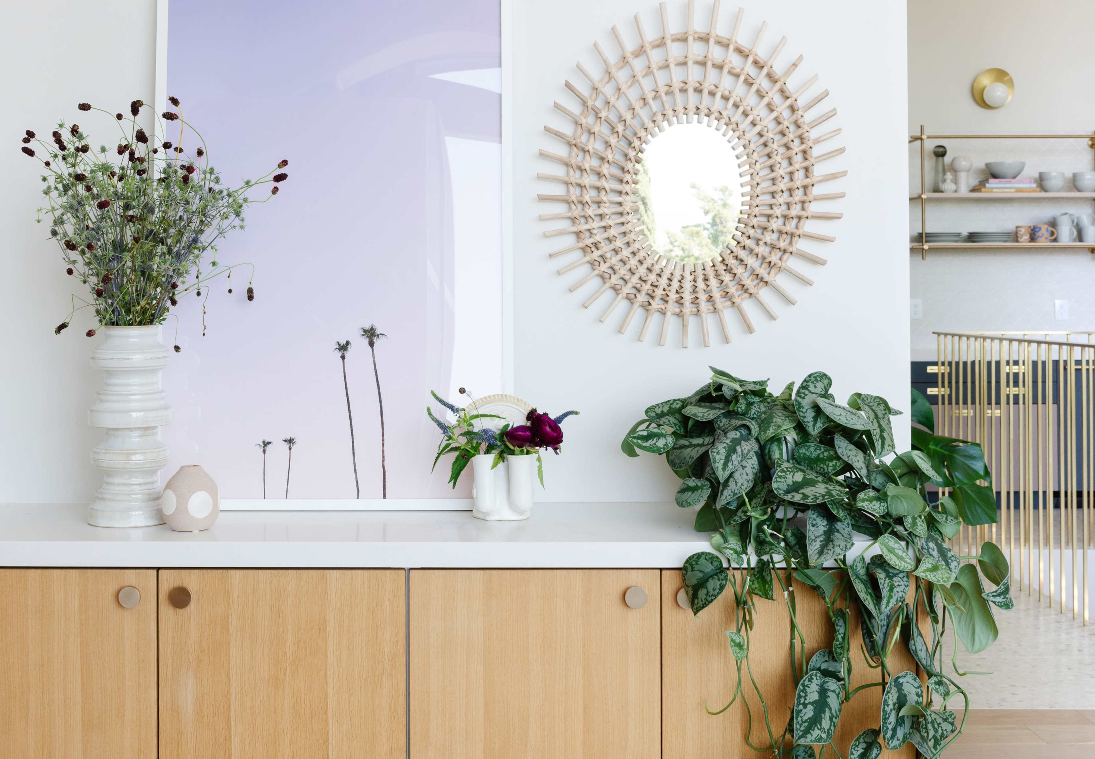 A modern interior features a wooden cabinet topped with decorative vases, a mirror, and a large leafy plant beside a framed artwork of palm trees.