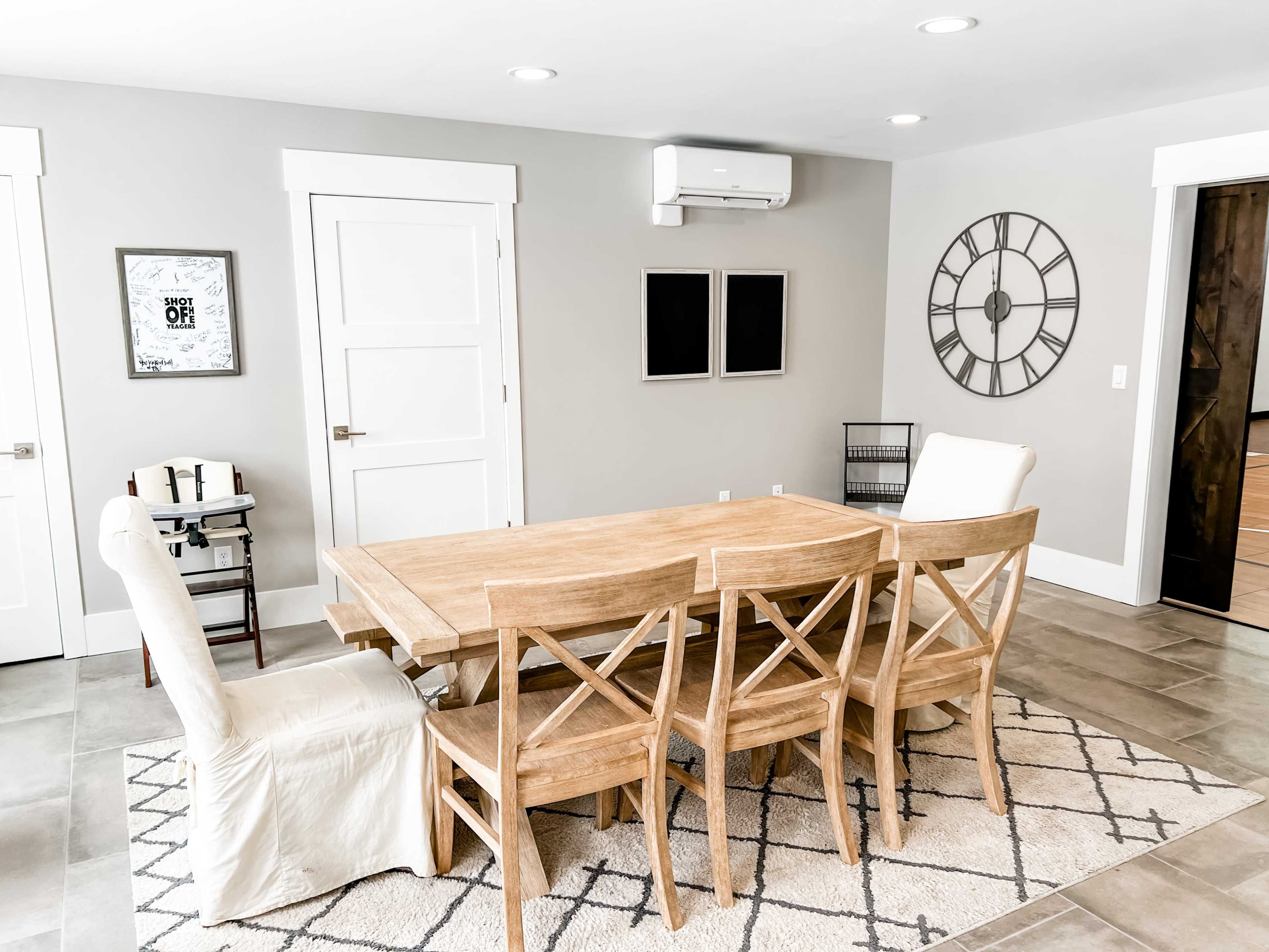 A dining area with a wooden table and chairs arranged on a patterned rug, along with a wall clock and several framed pieces on the walls.