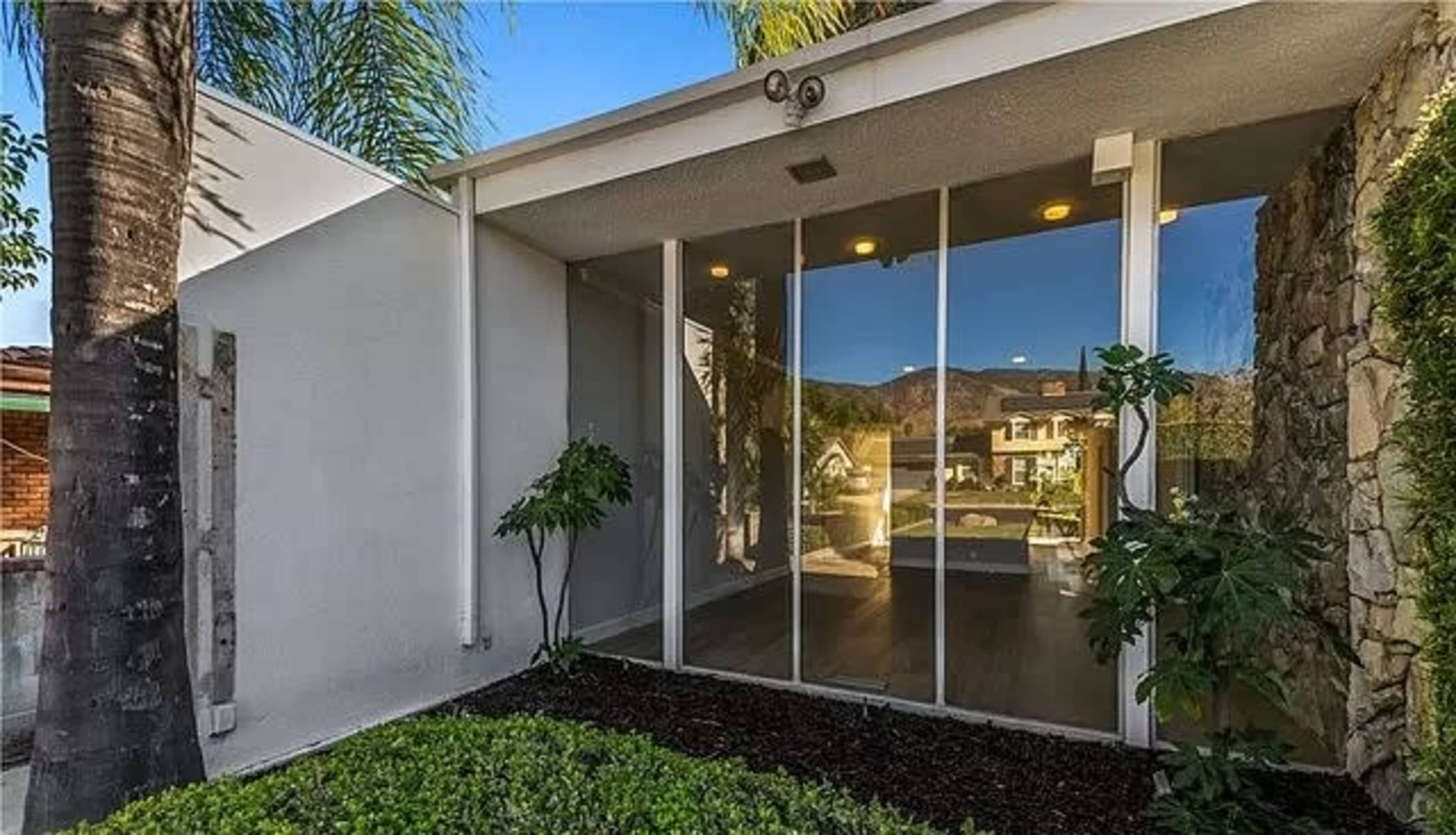 A modern home entrance with large glass windows, surrounded by lush greenery and palm trees.