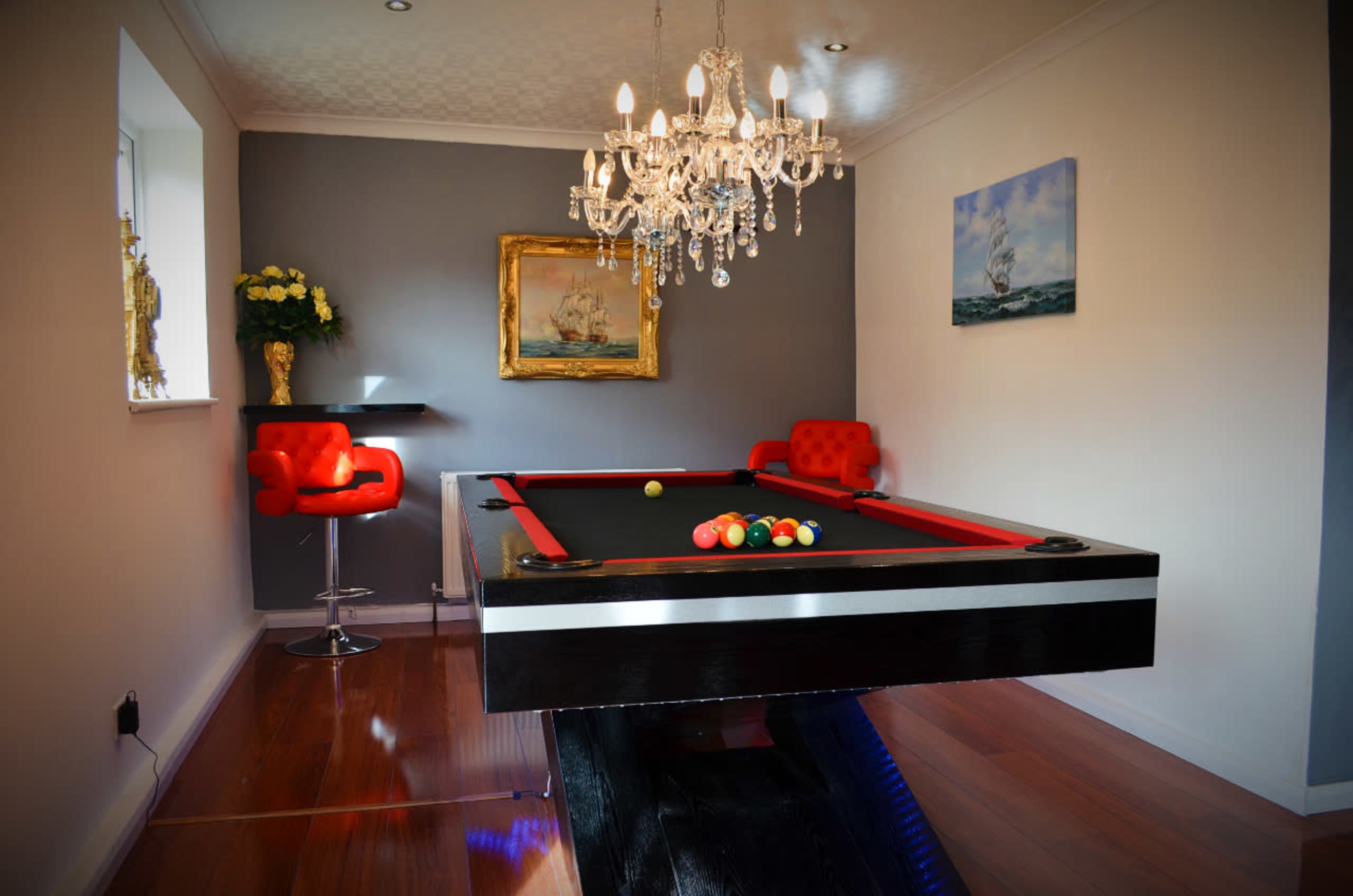 A modern game room featuring a billiard table with colorful balls, a chandelier, red chairs, and a decorative painting on the wall.