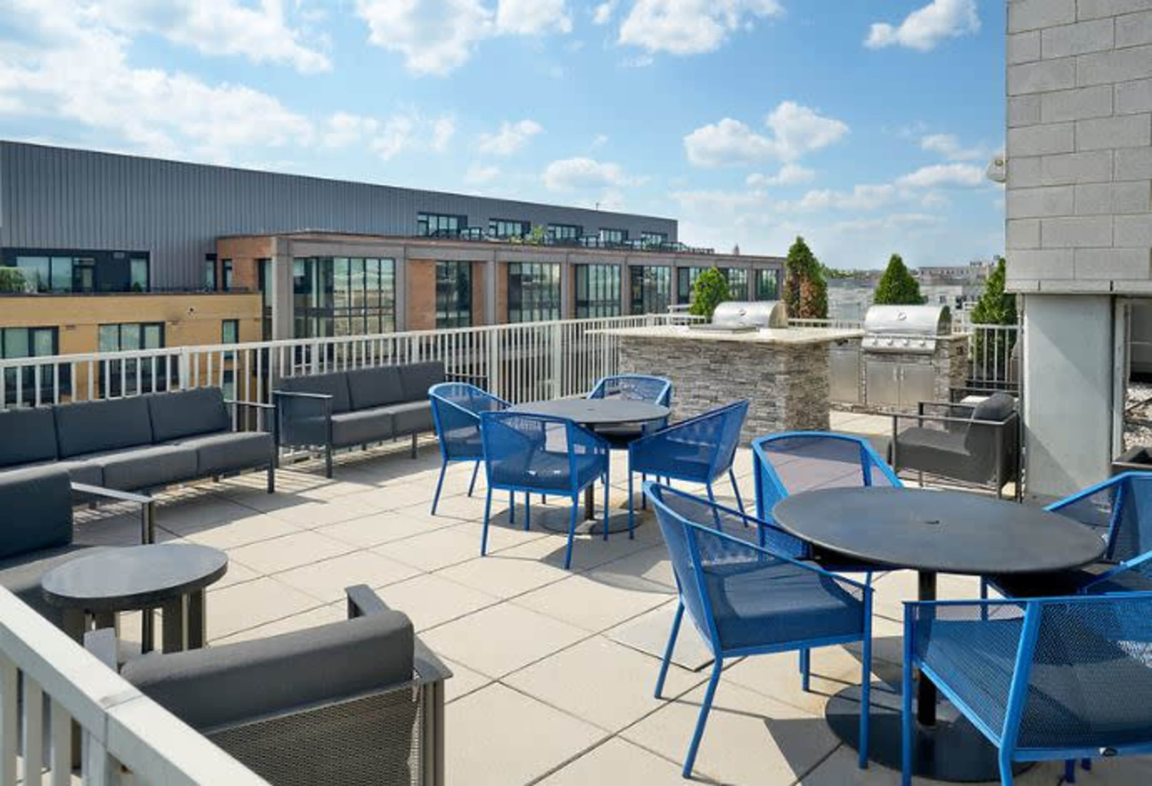 A rooftop patio with blue chairs and tables, surrounded by modern buildings under a partly cloudy sky.
