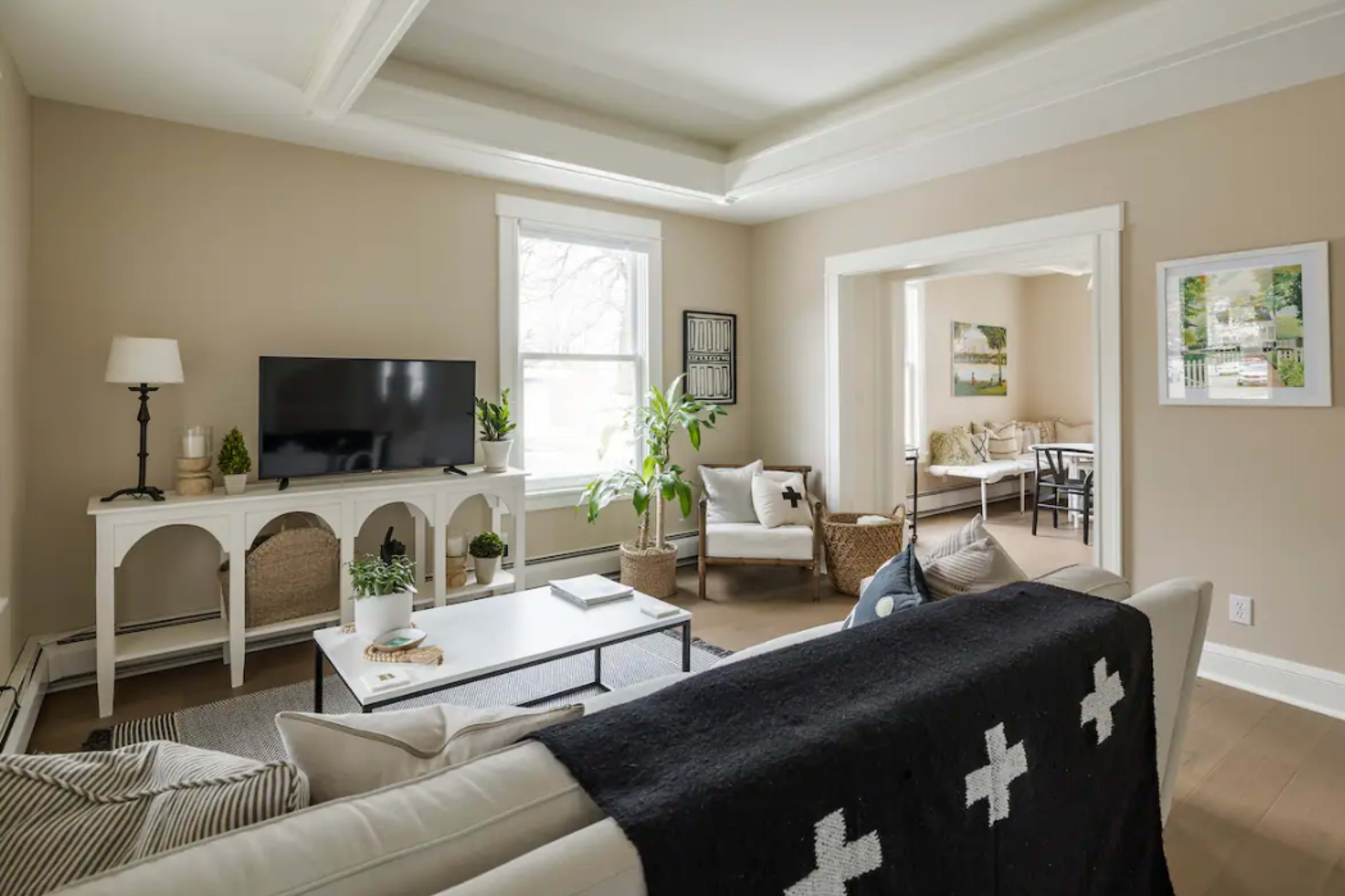 A light-filled living room features a television, a white console table, and a mix of seating arrangements with plants and decorative elements.