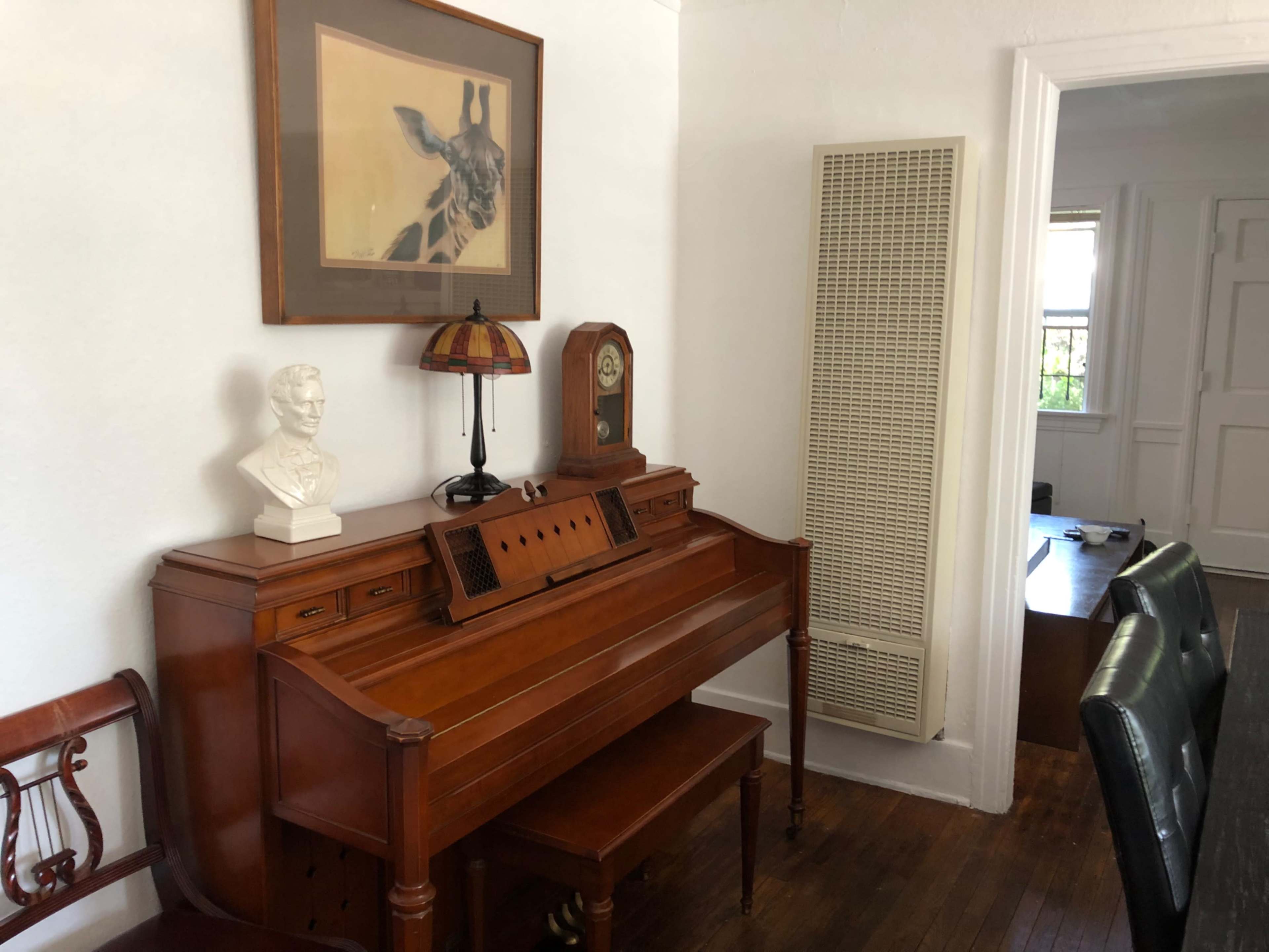 A wooden piano with a matching bench is positioned against a white wall, accompanied by a bust sculpture and a vintage lamp, while a heater stands nearby.