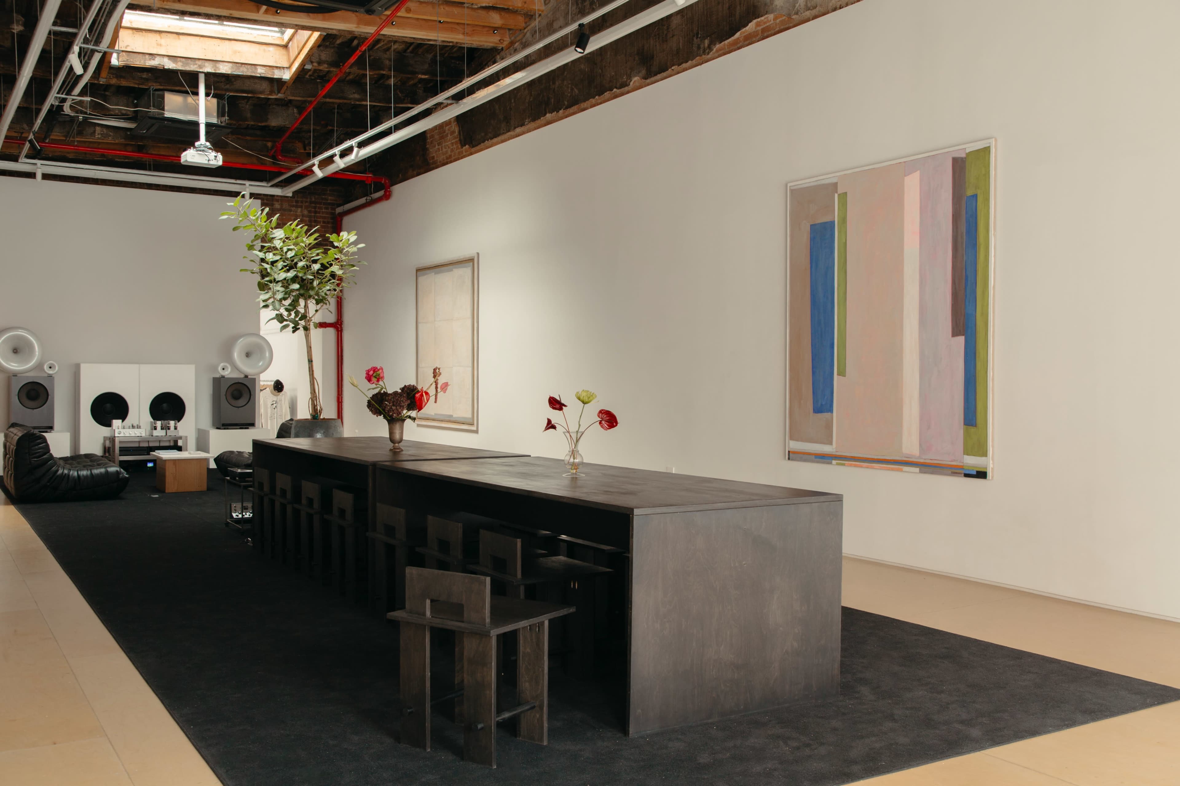 The image shows a modern interior space with a long, dark wooden table surrounded by minimalist chairs, a large piece of abstract art on the wall, and speakers and a black sectional sofa in the background.