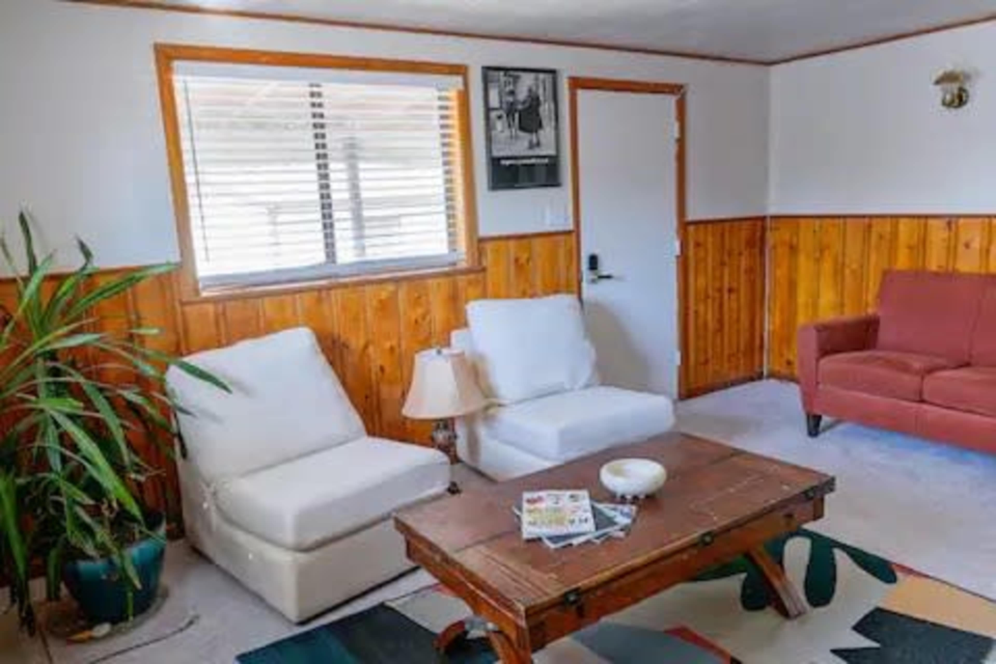 The image depicts a living room with two white armchairs, a red sofa, a wooden coffee table, and wooden paneling on the walls.