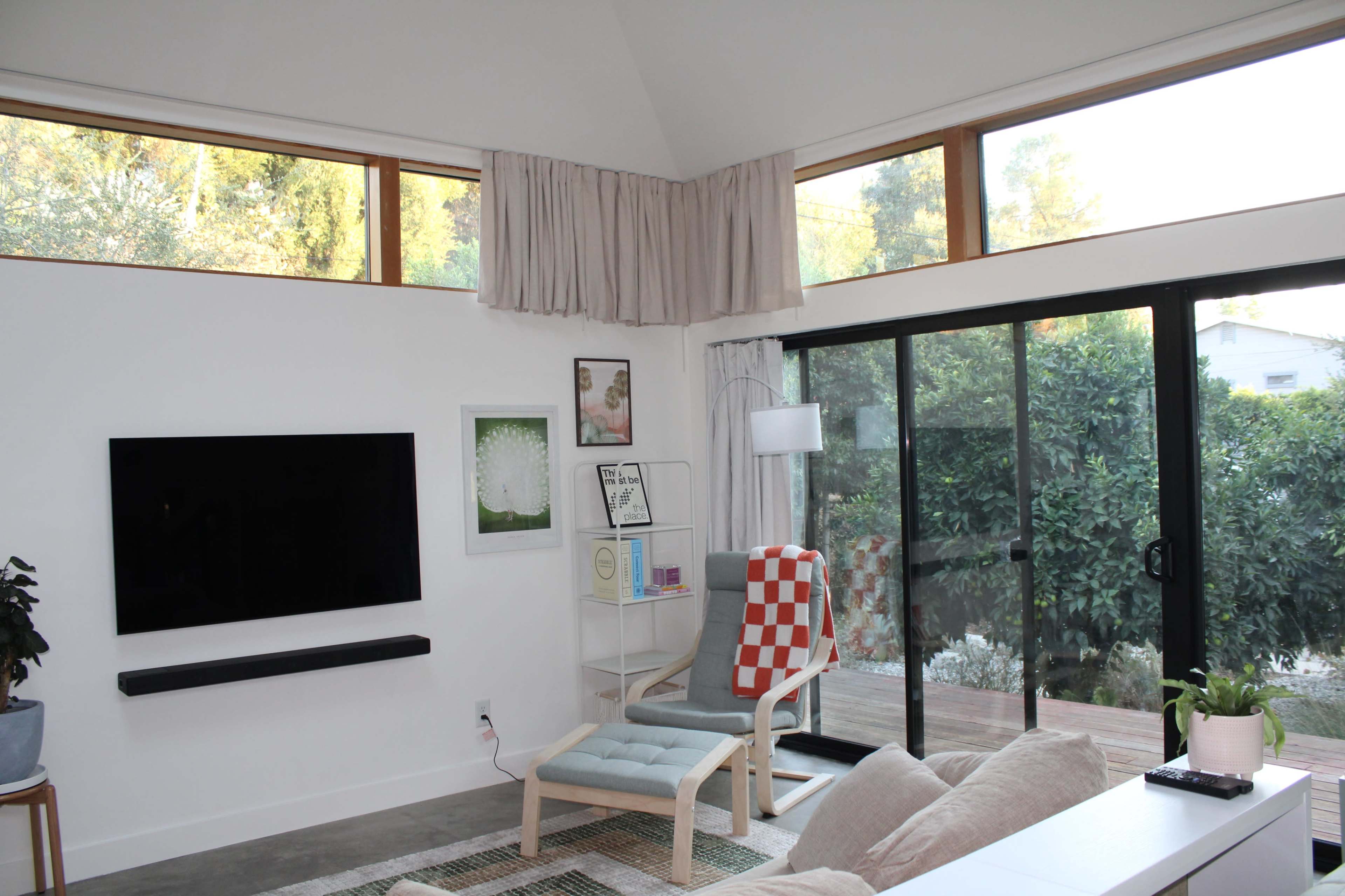 The image shows a modern living room with large windows, a wall-mounted TV, a light blue chair, and a sliding glass door leading to an outdoor space.